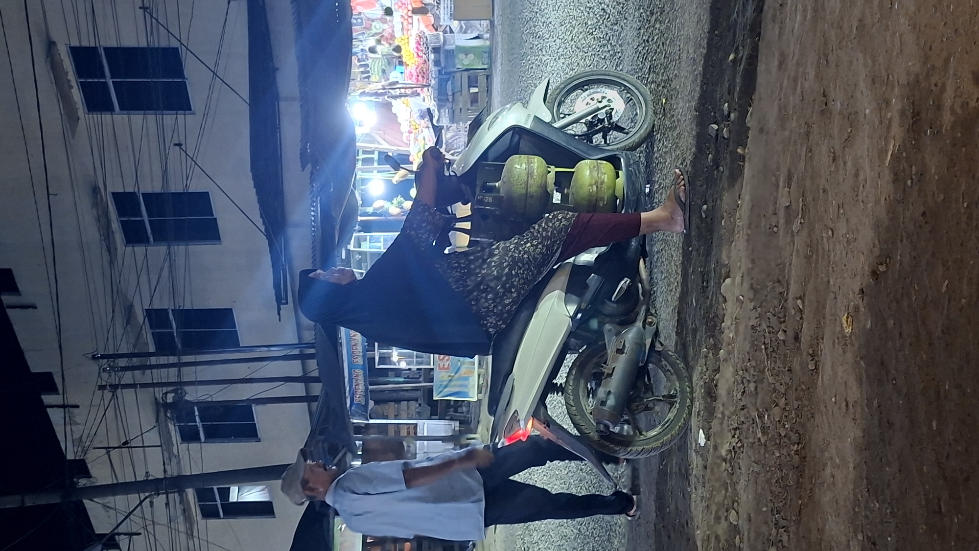 The image depicts an outdoor scene at night in Keudegeudong, Indonesia. A woman wearing a dark hijab and patterned clothing is riding a white scooter. Strapped to the side of the scooter are two green propane gas cylinders. Her legs are clad in maroon trousers, and she wears brown flip-flops. In the background, a man in a light blue shirt and dark pants, wearing a light-colored cap, stands near the scooter. Behind the people and scooter, a building with multiple dark-paned windows is visible. A street with overhead electrical wires runs across the frame. A market stall with bright lights displaying fruits and goods is also present. The ground is a mixture of gravel and dirt. The overall lighting suggests it is after dark, with artificial light sources illuminating the area.