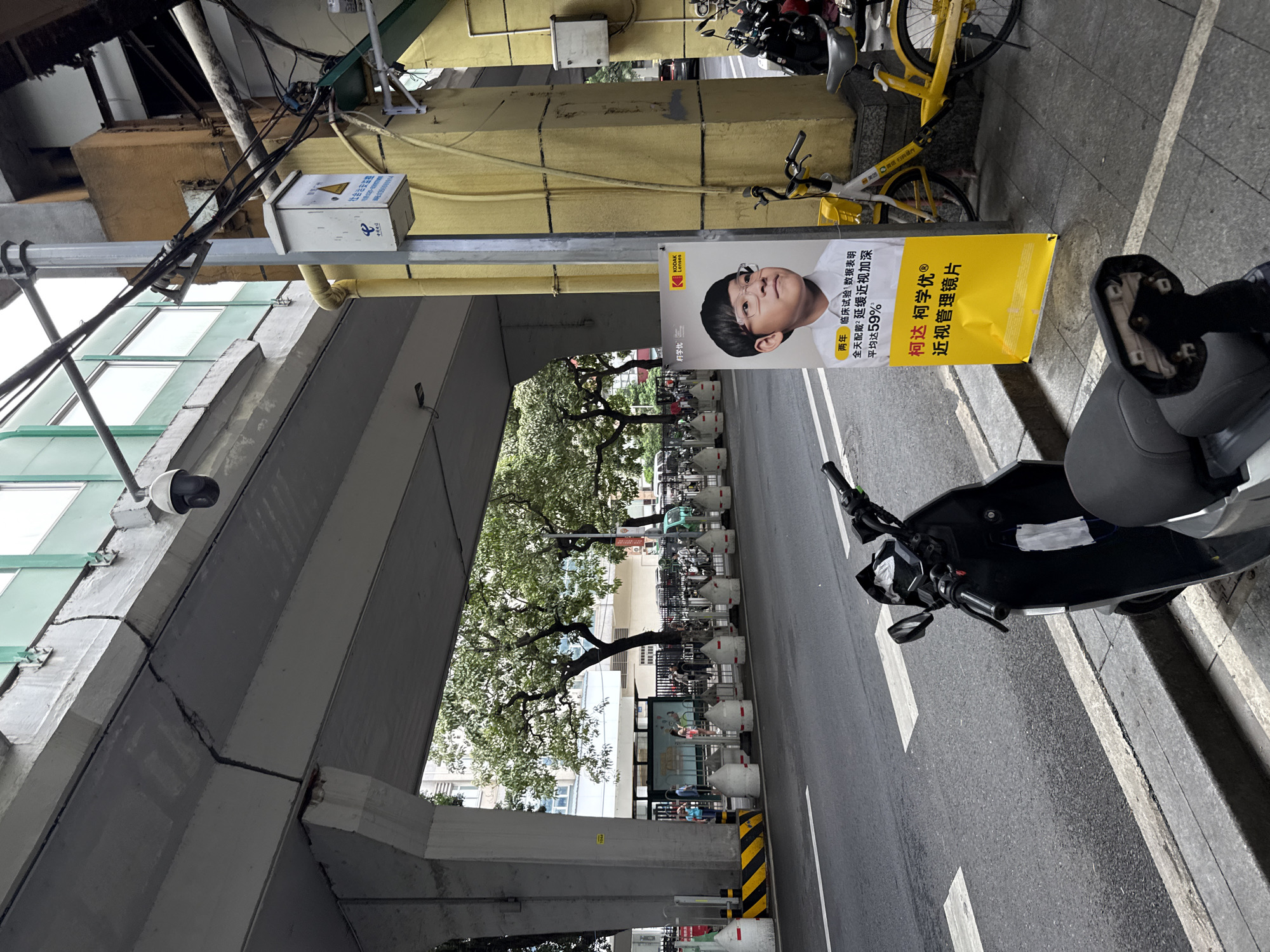 An outdoor, street-level, medium-shot photograph captures a section of an overpass in Guangzhou's Yuexiu District, China. The concrete structure of the overpass dominates the upper portion of the image, showing signs of weathering and a prominent crack. Below the overpass, a street runs parallel to the sidewalk. Several white, concrete bollards line the edge of the sidewalk, separating it from the road. A black electric scooter is parked on the sidewalk in the foreground, with a white surgical mask hanging from its handlebars.
To the right of the scooter, a tall, yellow advertising poster is propped against a concrete pillar. The poster features a portrait of a young boy wearing glasses, with Chinese text overlaying the image. The text appears to be an advertisement for Kodak Lenses, promoting myopia control. To the right of the poster, a yellow bicycle, also a shared bike, is visible. Beyond the sidewalk and to the left, a row of parked bicycles or scooters is partially obscured by the overpass pillars. People are faintly visible in the background, near what appears to be a bus stop or waiting area. The overall scene depicts an urban environment with typical street furniture and infrastructure.