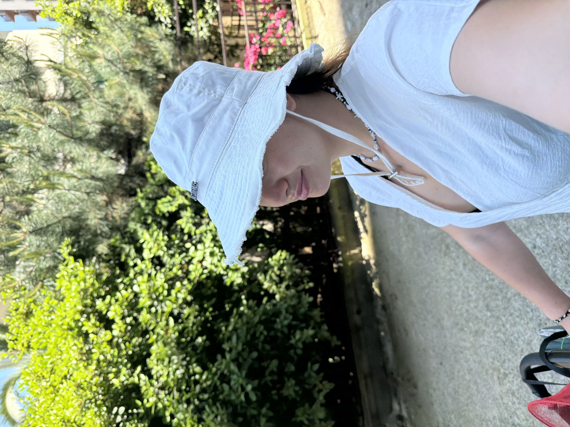 An outdoor, eye-level shot, likely taken in Sochi, Russia, captures a person wearing a white bucket hat and a white crinkled long-sleeved top. The image is tilted, presenting a dynamic perspective. The person's face is partially obscured by the hat's brim, but their lips are visible in a slight, serene smile as they look downwards and to the right. A beaded necklace, featuring blue, white, and shell-like elements including a star, adorns their neck.
The white bucket hat has a small silver-colored metal tag on its side, clearly displaying the text "JACQUEMUS". The person also wears a black and white beaded bracelet on their right wrist. Partially visible in the lower right corner are what appear to be the handlebars of a bicycle or stroller, with sunglasses hanging from them, and a section of a red mesh bag.
The background is filled with lush green foliage, including various bushes and trees, some appearing to be pines. In the middle ground, on the right side of the frame, vibrant pink flowers add a splash of color. The ground beneath the person is a light-colored concrete or paved surface. The bright lighting suggests it is daytime, likely sunny, indicating pleasant weather for an outdoor activity. The overall scene conveys a casual, relaxed atmosphere.