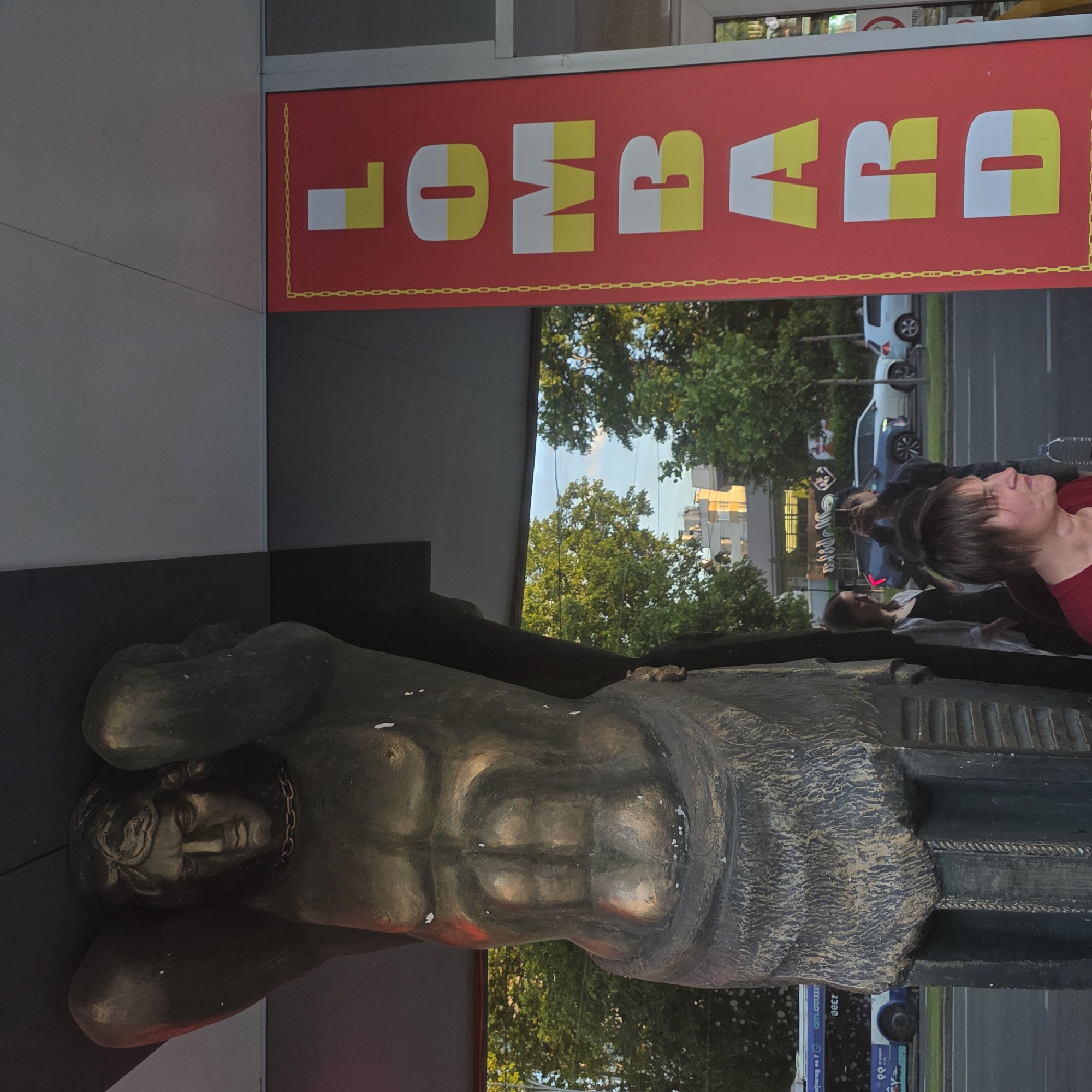 A bronze statue of Atlas, a muscular male figure with his arms raised and supporting a dark structure above, stands prominently in the foreground. The statue is detailed, showing defined musculature on the abdomen and chest, and a stern facial expression. A chain is visible around the statue's neck. To the right of the statue, a large red sign with yellow and white lettering spells out "LOMBARD" vertically. The sign is bordered by a yellow chain pattern. In the background, trees with green foliage line a street where a blue bus and several cars are visible. The sky above appears clear. Several individuals are present in the scene; one woman in a red shirt is in the immediate foreground, looking to the right. Behind her, a woman in a white shirt and black top stands, and a person behind her is holding up a phone, seemingly taking a picture.