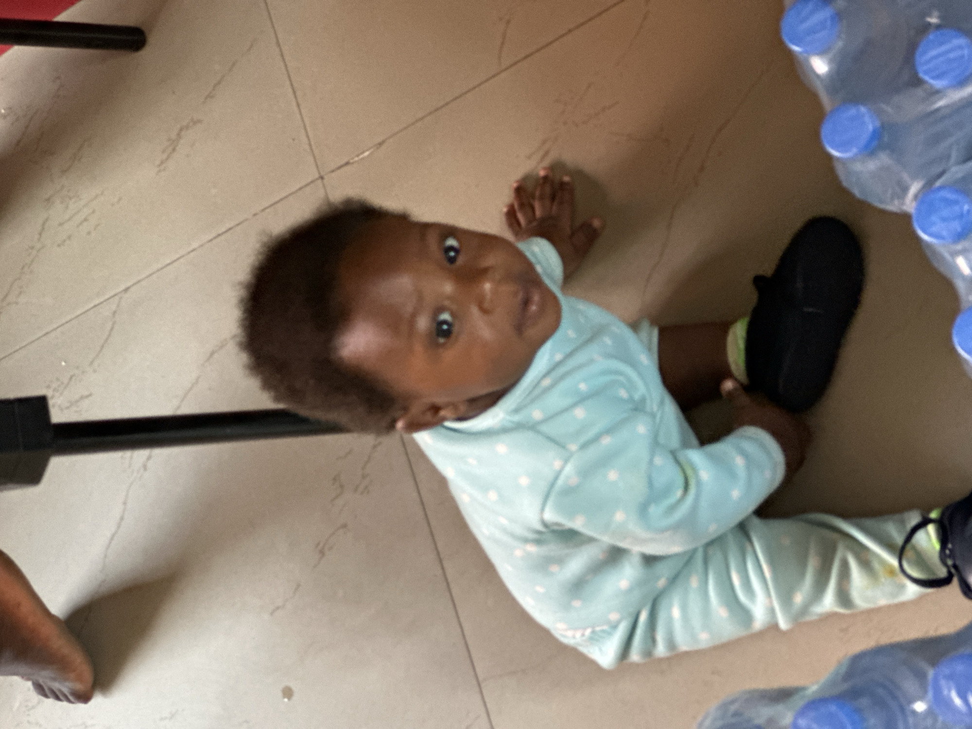 A low-angle, close-up shot features a young child, likely an infant or toddler, sitting on a light-colored, patterned tiled floor in an indoor setting. The child has dark skin and short, dark, curly hair, and is looking directly at the camera with wide, curious eyes. They are dressed in a light blue, long-sleeved onesie patterned with small white polka dots. A black shoe is worn on the child's right foot, while the left foot is bare.
Clustered on the right side and extending towards the bottom of the frame are several large, clear plastic water bottles, each topped with a distinct blue cap. On the far left, a dark, horizontal bar (possibly part of furniture or an object) is visible, along with a portion of an adult's bare foot. The tiled floor exhibits subtle marbling and visible grout lines. A small, yellowish stain is noticeable on the lower left leg area of the child's onesie. The scene suggests a casual, everyday moment in an indoor environment, likely a home in Jos, Nigeria, given the provided context. No visible text is present.