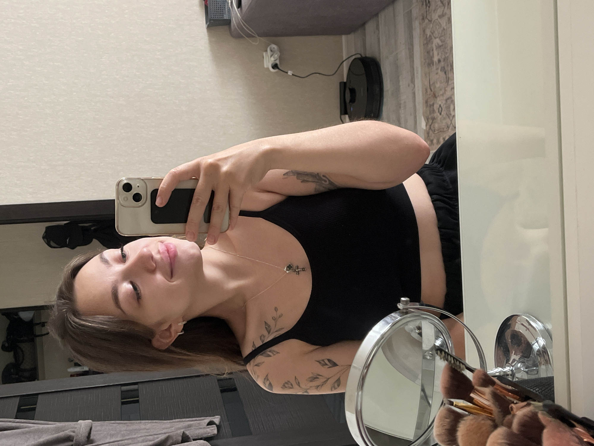 A young woman with long brown hair and tattoos on her arm is taking a selfie in a mirror. She is wearing a black tank top and black sweatpants. She is holding a smartphone in her right hand, and her left hand is resting on her hip. In the foreground, there is a makeup mirror and a collection of makeup brushes. In the background, there is a closet, a robot vacuum cleaner, and a sofa. The image appears to be taken in a bedroom or dressing area in an apartment in the city of Chaikovsky, Russia. The lighting is natural and soft, creating a relaxed and intimate atmosphere.