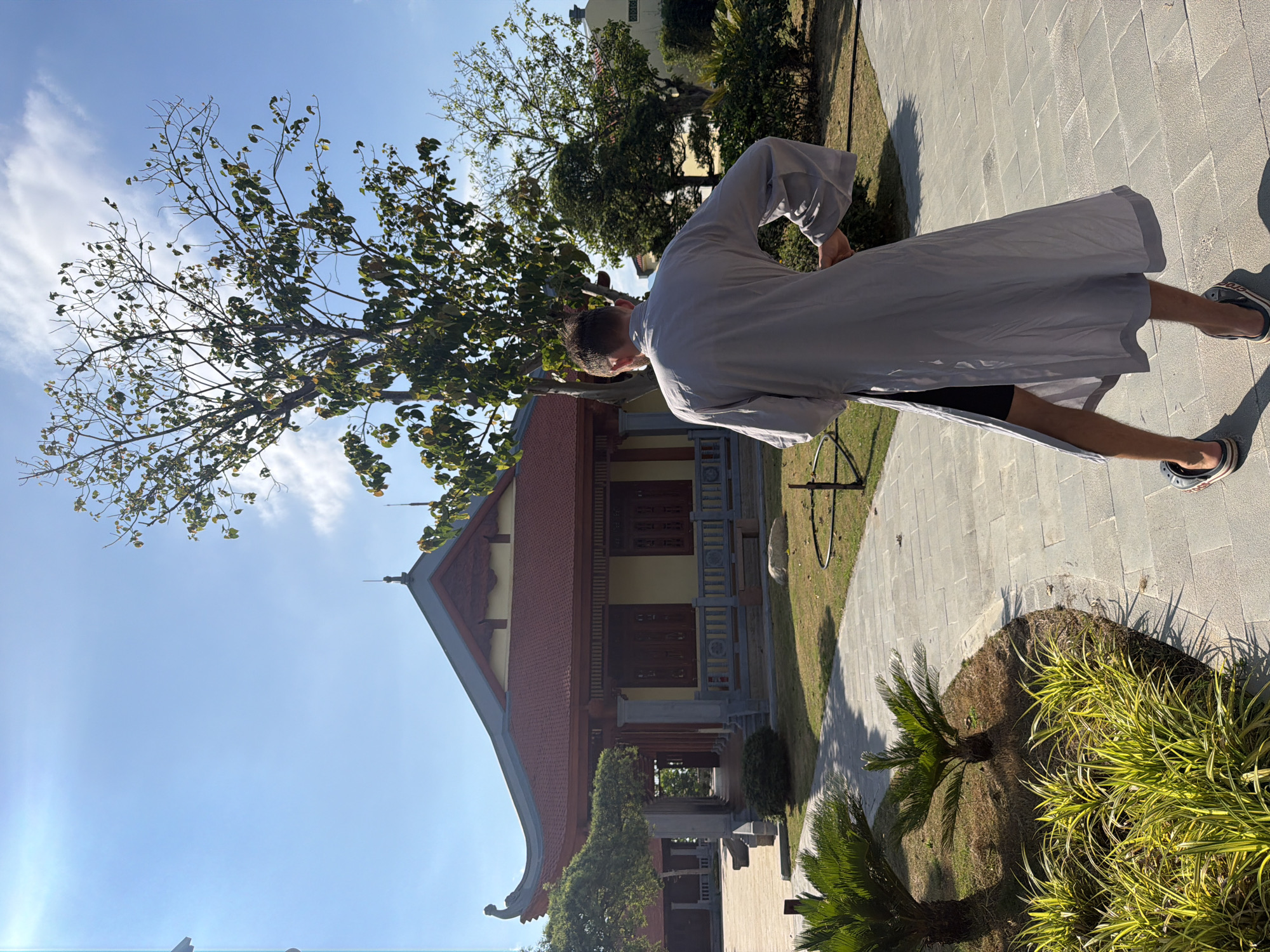 A medium shot of a person wearing a grey robe standing outdoors in Nam Nha Trang Ward, Vietnam. The person's head is turned towards the left, and they are wearing sandals. Behind the person is a building with a red roof. A tree with thin branches is to the left of the building. To the right of the person, there is a paved walkway and a patch of grass with plants. The sky is blue with some clouds.