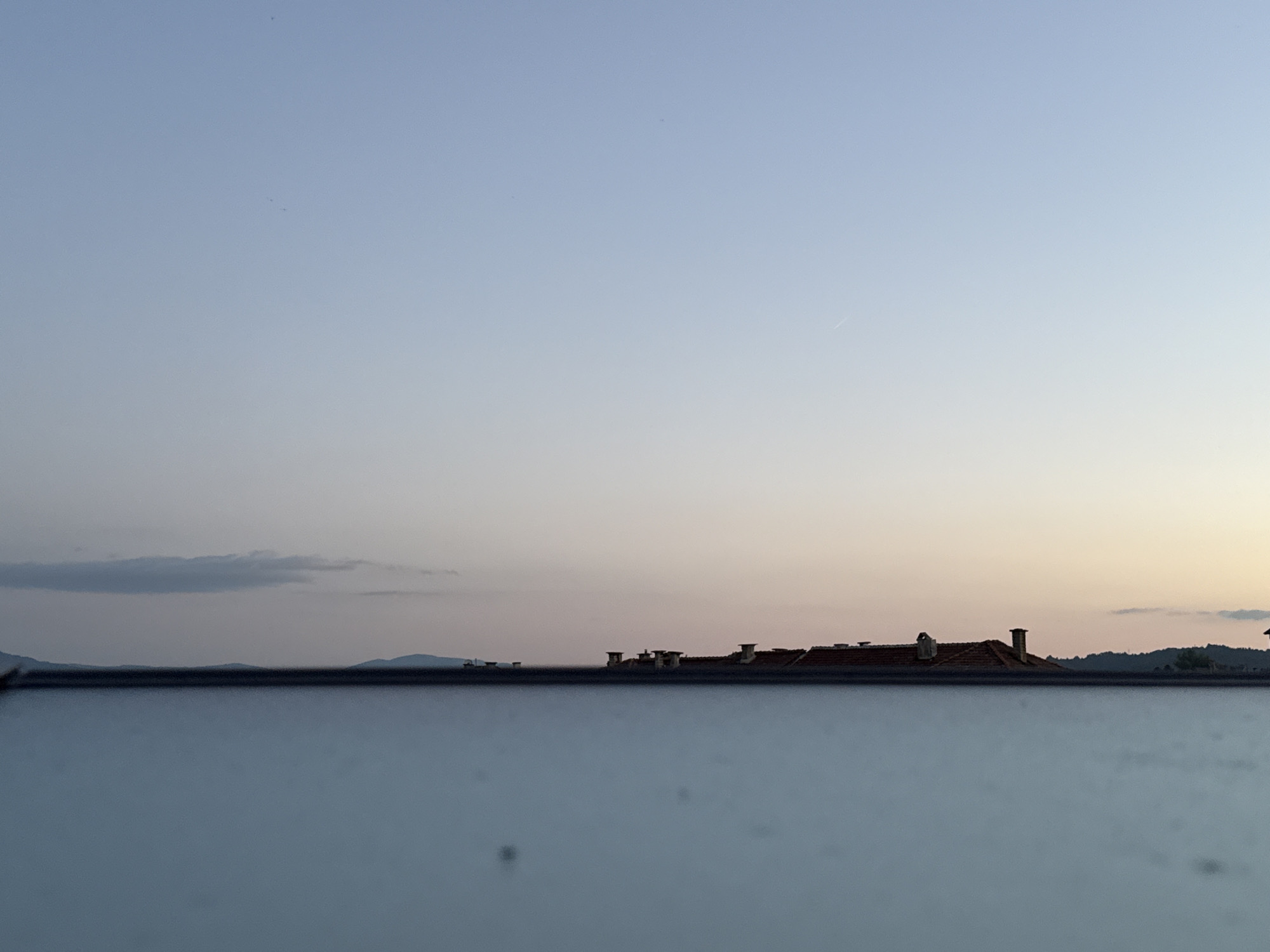 The image shows a broad, horizontal scene composed of a clear, pale blue sky gradually transitioning to a soft pinkish-orange hue near the horizon. The lower half of the image is dominated by a smooth, reflective surface, likely water or glass, which mirrors the sky and the scene above.
In the middle ground, a dark, sharp horizon line separates the sky from the reflective surface. Along this horizon, silhouetted against the lighter sky, are rolling hills or mountains. Dotted along the top of these hills are the indistinct shapes of buildings with visible rooftops and chimneys. These structures appear to be residential, with terracotta-tiled roofs. The overall impression is a tranquil, possibly dusk or dawn, setting.
Based on the architectural style and the landscape, this scene is likely located in or near Blagoevgrad, Bulgaria, a city known for its proximity to mountainous terrain and its characteristic Balkan architecture. The specific location within Blagoevgrad is not discernible from the image alone, but the presence of distant hills and scattered buildings suggests an edge-of-city or semi-rural environment. There are no people or discernable actions visible in the image. The environment is calm and serene.