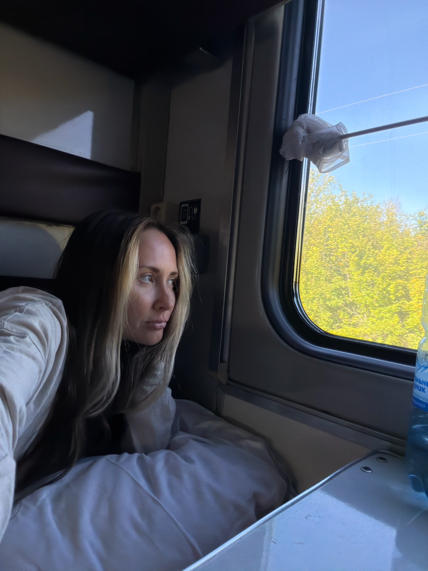 A woman with long, blonde-streaked brown hair is lying down on a bed inside a train compartment. She is looking out of the window to her right. Outside the window, green trees are visible against a clear blue sky. A silver pole with a white cloth attached to the end is extending from inside the compartment towards the window. A blue plastic bottle of water is visible on a surface to the right of the window. The interior of the train compartment is dimly lit, with some light reflecting on the walls and a table. The woman is wearing a light-colored long-sleeved shirt. This scene is set within a train compartment traveling through the city of Lapshinskaya, Russia.