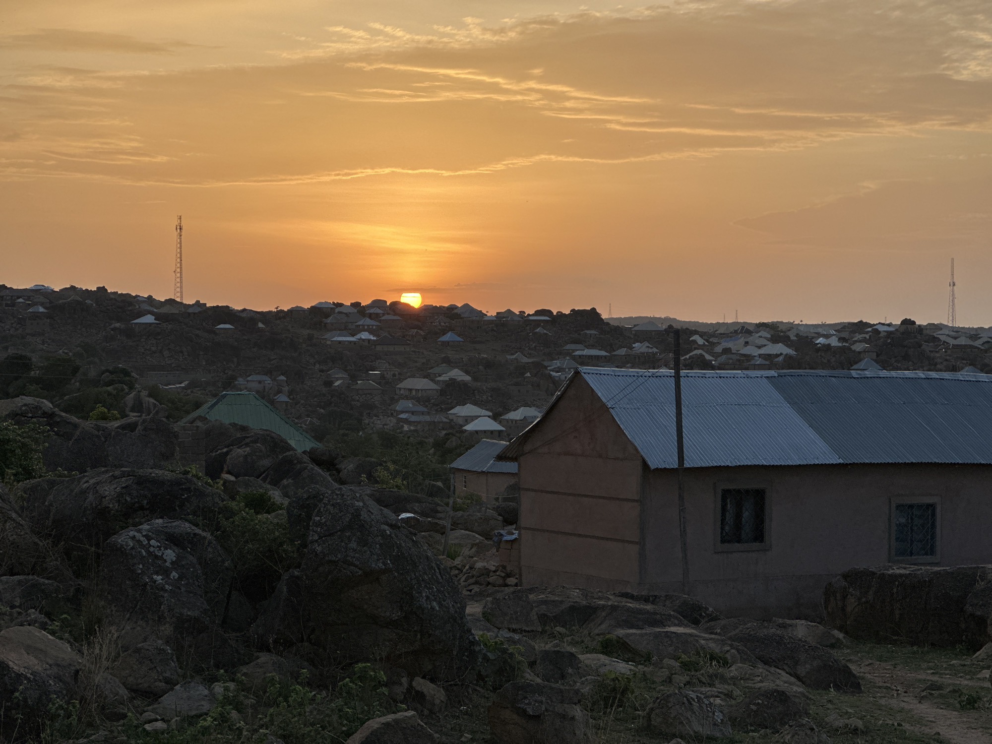 The image shows a sunset view of a residential area in Kabong, Nigeria. The sky is a gradient of orange and yellow. The sun is partially obscured by a rocky ridge dotted with houses. In the foreground, there are large boulders and a house with a corrugated metal roof and two visible windows. A communication tower is visible in the distance on the left.