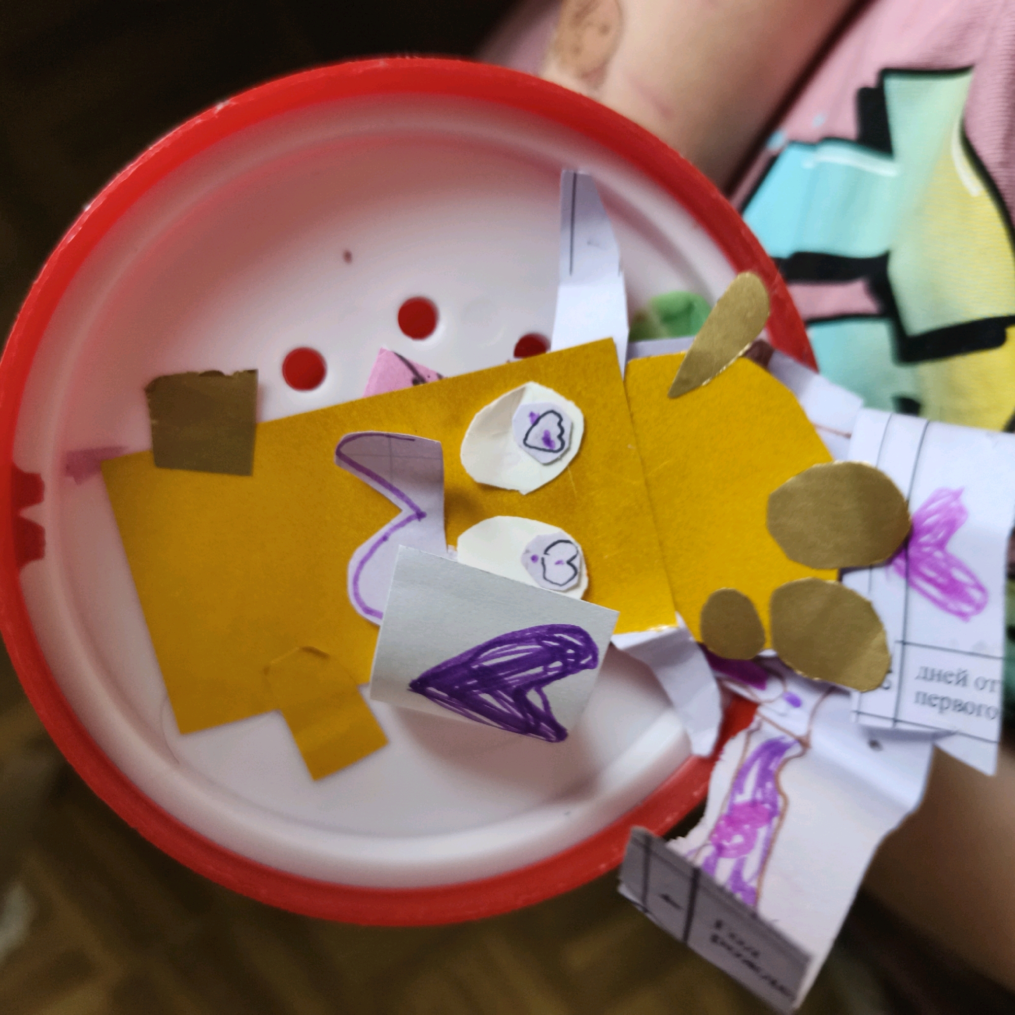 The image shows a close-up of a red-rimmed white plastic container, likely a lid, filled with various colorful paper cutouts and drawings. These appear to be elements of a craft project. Among the items are pieces of yellow, pink, and white paper, some with drawn purple markings and shapes. There are also small, oval-shaped brown paper cutouts that resemble eyes or buttons. The background consists of a person's arm and shoulder wearing a brightly colored t-shirt with abstract designs in pink, blue, and yellow, suggesting a domestic or creative environment. The image is not specific to any location within Chaikovsky, Russia, and focuses solely on the contents of the container.