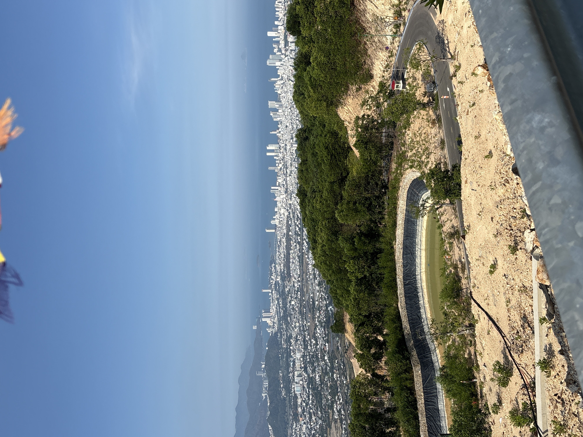 A high-angle view from a mountainous area overlooking the city of Nam Nha Trang Ward, Vietnam. The city sprawls along the coast, a dense collection of white buildings under a clear blue sky. Between the viewpoint and the city lies a stretch of ocean, its surface smooth and reflective. A road winds along the mountainside, with at least one vehicle on it. The terrain is arid and rocky, with patches of green vegetation clinging to the slopes. In the bottom left of the frame, a railing or part of a structure is visible.