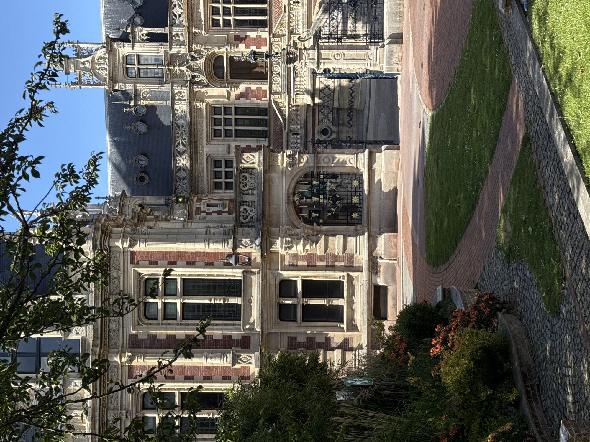 This image captures an ornate historic building, likely a grand château or palace, set against a clear blue sky on a bright, sunny day. The scene suggests a well-maintained historical site or museum, with landscaped grounds in the foreground.
The central subject is the multi-story building, distinguished by its intricate architecture. The facade features a striking combination of light-colored stone and reddish-brown brick arranged in decorative patterns. Numerous windows of various sizes and shapes, some with multiple panes, are visible across the building's face. The structure is adorned with elaborate stone carvings, balustrades, and decorative elements, giving it a rich, gothic or renaissance revival appearance. A prominent dark-tiled roof with several dormer windows and smaller circular openings adds to its grandeur. Below the main windows, decorative metal grates, one featuring star-like motifs, are visible.
In the foreground, lush green foliage from trees and shrubs partially frames the building on the left and bottom right. These plants cast long shadows across the facade and the ground, indicating the strong sunlight. To the right, landscaped grounds feature a winding pathway made of reddish brick pavers, contrasting with a cobblestone path and sections of neatly mown grass. Flowering plants and hedges line the paths and the base of the building.
The bright lighting suggests it is daytime, with clear weather conditions. No people are visible, and no specific activity or event is taking place, presenting a serene and static view of the architectural masterpiece and its surroundings. A small, partially obscured green sign with white, illegible text is visible among the foliage on the right side.