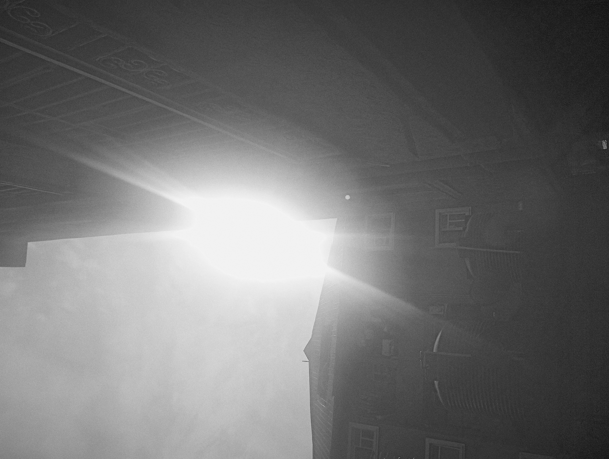 The image, in black and white, depicts a bright light source (possibly the sun) shining on a building. The building appears to be multi-story with windows and balconies visible. The perspective is angled, looking upward at the building and light. The location is unspecified and cannot be determined solely from this image.