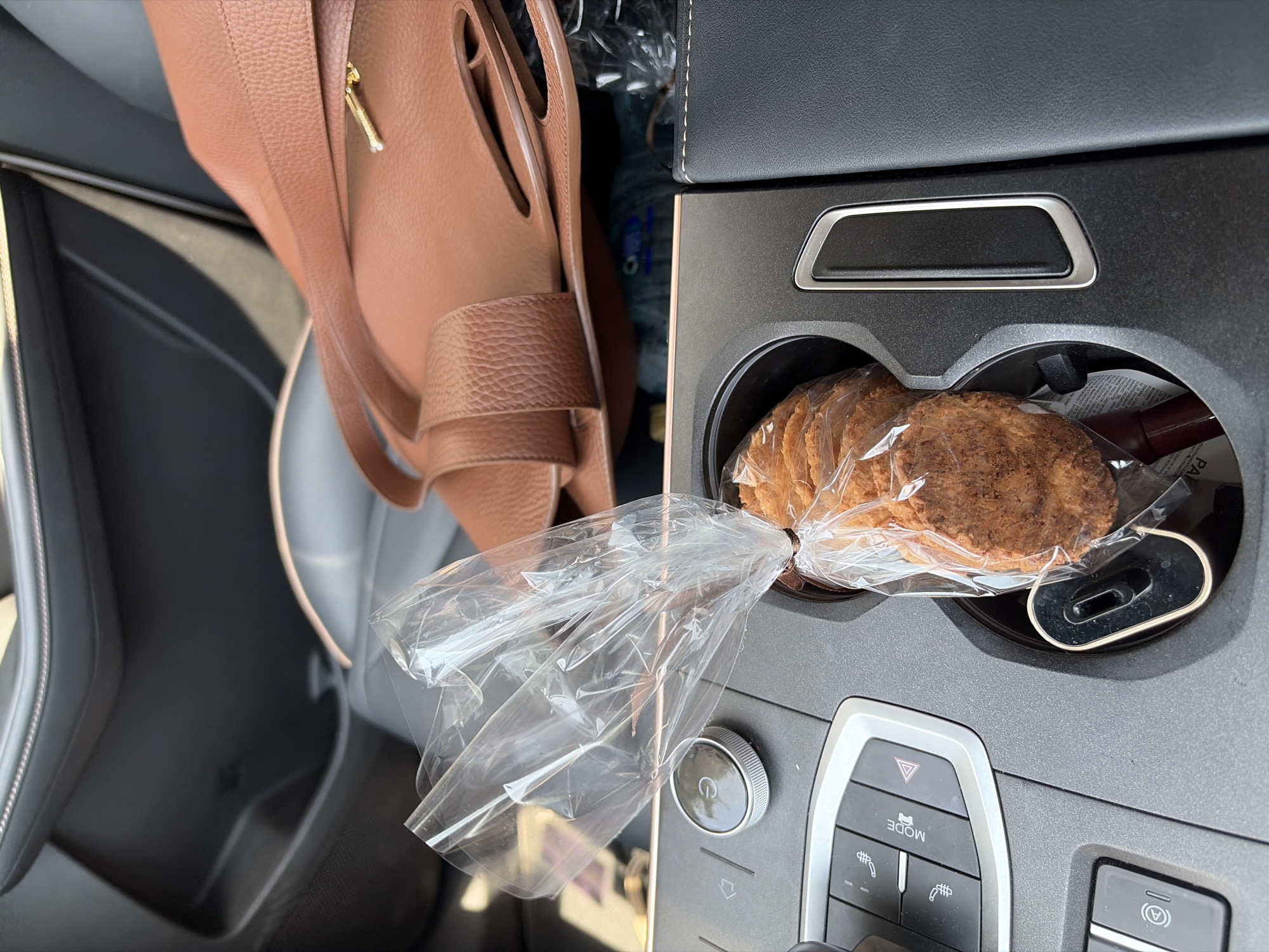 The image, likely taken from within a car, shows a tan leather handbag hanging near the driver's seat, alongside a bag of cookies placed in the car's cupholder. The interior elements visible suggest the scene is in a car, including the seat, dashboard, cupholder with cookies, and a plastic bag. The specific location within Puebla, Mexico, is not identifiable from the image.