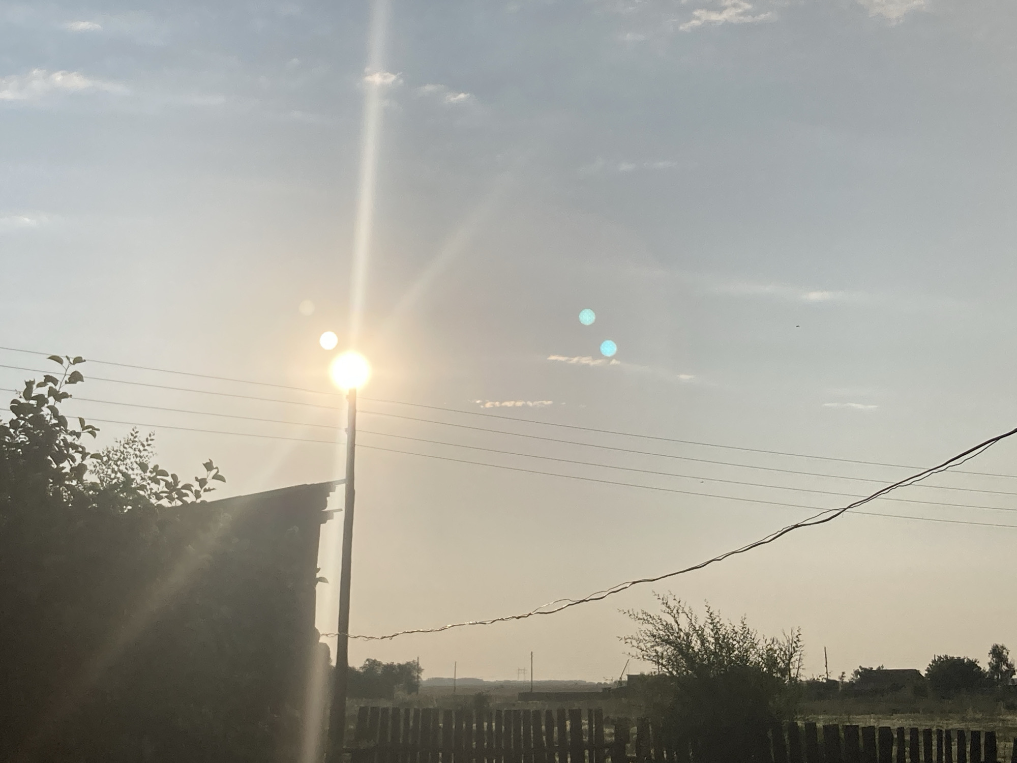 This is a horizontal, full shot image of a rural landscape in Rakity, Russia, taken during the daytime. The sun is the dominant subject, positioned in the upper left quadrant of the image, emitting bright rays that create lens flares and cause overexposure in the surrounding sky. The sky itself is a hazy blue, with scattered white clouds.
In the foreground, on the left, is a dark, dilapidated structure, possibly a shed or an old building, partially obscured by dense green foliage. To the right of the structure is a tall, thin pole, likely a lamppost, with a bright light fixture at its top, also contributing to the sun's glare. Several power lines are strung across the frame, running horizontally from left to right.
The middle ground features a wooden fence made of vertical posts, behind which a low horizon is visible. This horizon line is dotted with trees and some indistinct structures, suggesting a small village or rural settlement. The overall atmosphere is serene and quiet, with the bright sunlight casting a warm, hazy glow over the scene. The time of day appears to be late afternoon or early evening, given the angle of the sun and the hazy quality of the light. There are no people visible in the image.
