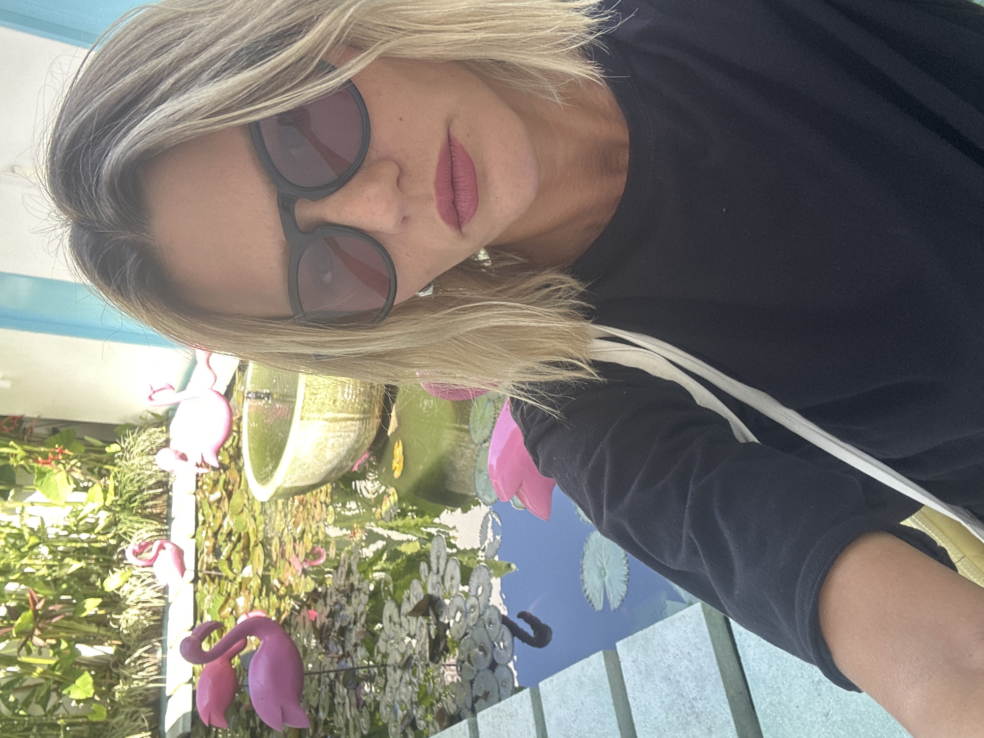 A woman wearing sunglasses, a black long-sleeved shirt with white stripes, and pink lipstick is facing the camera, with a partial view of a pond in the background. The pond features a tiered fountain, lily pads, and pink flamingo figurines scattered around the edge. The scene appears to be outdoors. The location is North Kuta, Indonesia.