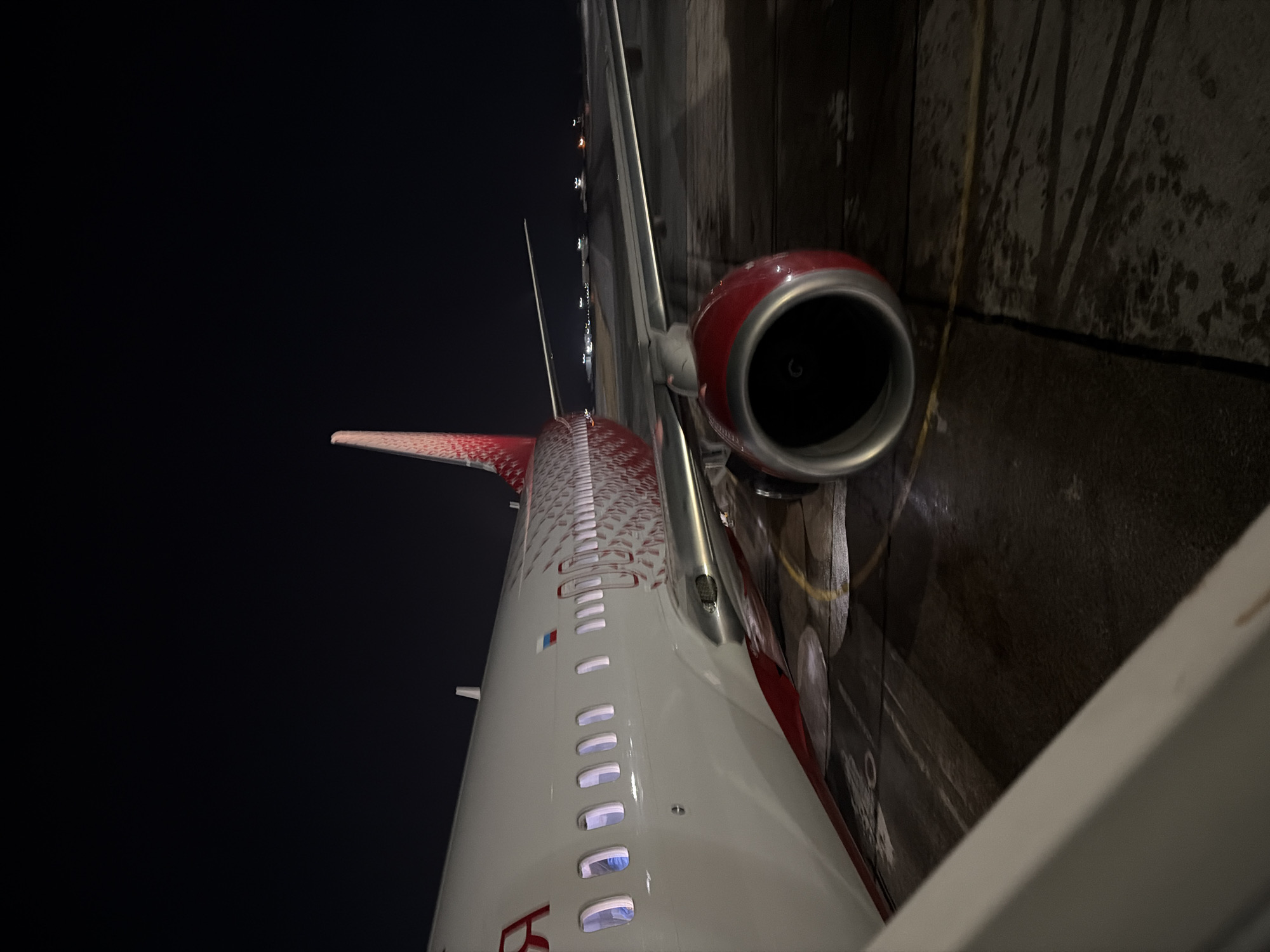 The image depicts the port side of a commercial passenger jet parked on a wet or icy airport tarmac at night, located in the vicinity of Сокол, Russia. The aircraft features a white fuselage with a distinctive red and orange stylized feather pattern on its tail fin and extending along the rear fuselage, characteristic of Rossiya Airlines. A small Russian flag is visible above the windows near the forward section, and the Cyrillic letter "Я" (Ya) is partially visible from the airline's name. The forward engine on the port wing has a red cowling with a dark intake. Multiple illuminated passenger windows are visible along the aircraft's side. Yellow ground markings are present on the dark, reflective tarmac. In the background, distant, blurred airport lights are visible under a dark night sky. A white structural element, likely part of a jet bridge or airstair, partially frames the lower left foreground. No individuals or specific actions are discernible.