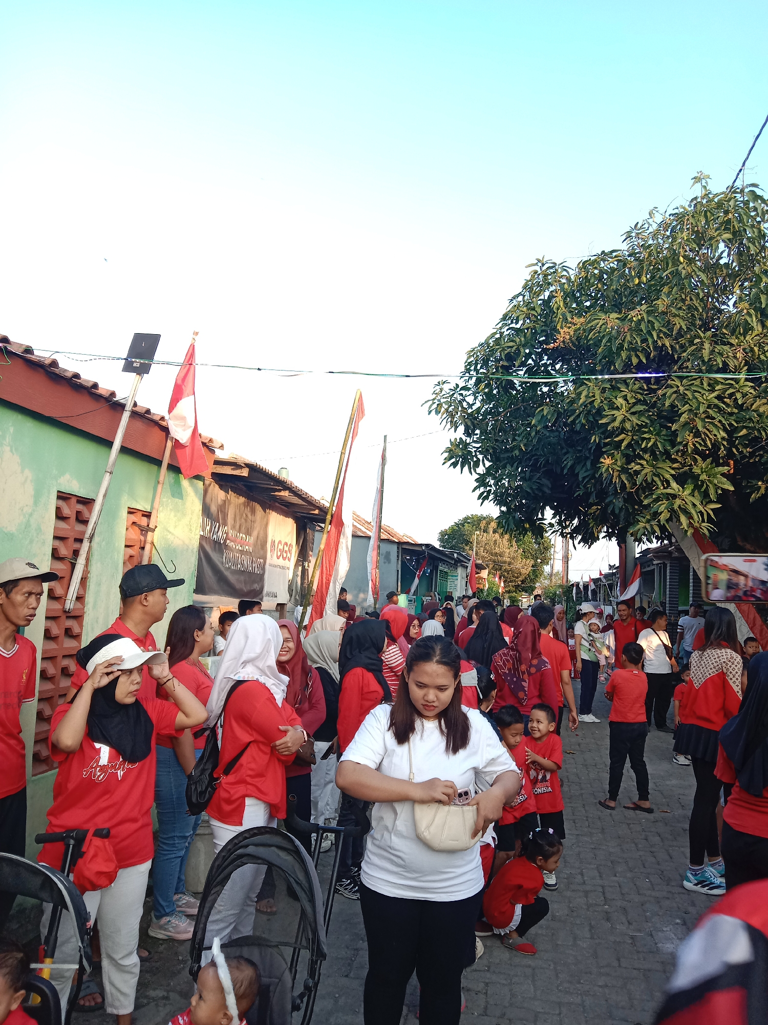 This is a street scene in Brangsong, Indonesia, possibly during a celebration given the prevalence of red and white attire and flags. A diverse group of people, including adults and children, are gathered along a narrow street lined with buildings and trees. Many individuals are wearing red t-shirts, and Indonesian flags are visible, some on poles and others held by people. The atmosphere appears lively and social, with people interacting and walking through the street. The buildings are simple, some with green painted walls and others with dark roofing. The ground is paved. The sky is a clear, bright blue, suggesting it's daytime, likely in the late afternoon given the angle of the sunlight. There are also baby strollers visible, indicating families are present.