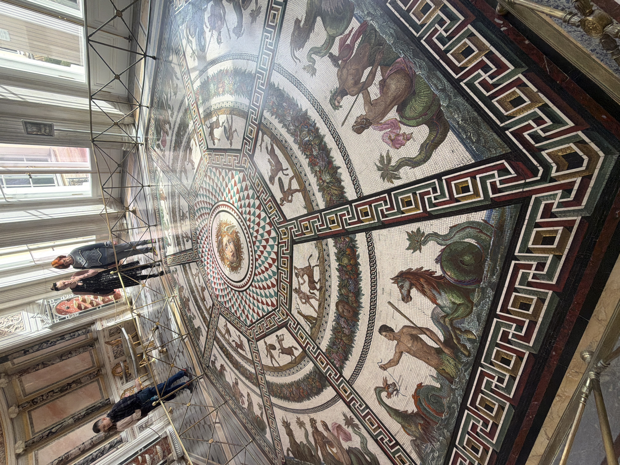 The image shows a large mosaic floor in what appears to be a grand hall with windows on the left side. Three individuals are visible from behind, standing on a platform that offers a view of the mosaic. The mosaic is ornate, featuring a central circular design with a face in the middle, surrounded by geometric patterns and depictions of animals and mythological creatures. The outermost border is a geometric pattern in shades of white, green, brown, and black. The mosaic spans a large area and appears to be made of small tiles. The hall has a classical architectural style. Given the details, this is likely the mosaic floor in the Hermitage Museum in Saint Petersburg, Russia.