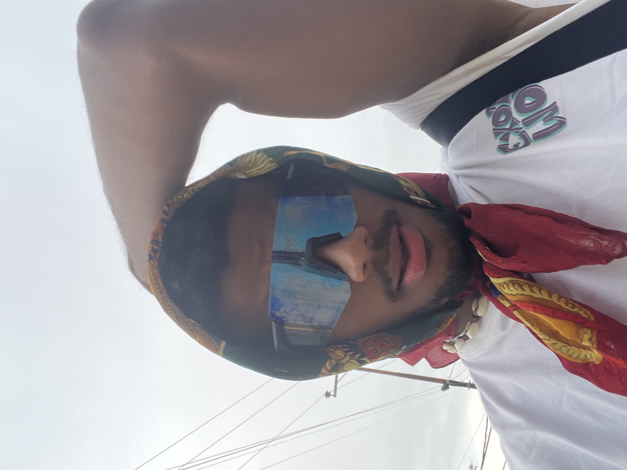 A man is captured in a selfie-style photo taken at Playa El Agua, Venezuela. He is wearing a patterned headscarf and blue-tinted sunglasses. A red bandana with gold accents is tied around his neck, and he wears a necklace made of seashells. He is wearing a white t-shirt with a black and white striped collar and a multicolored logo. Part of his arm is visible in the upper left corner of the frame. Wires and masts from boats can be seen in the background against a bright sky.