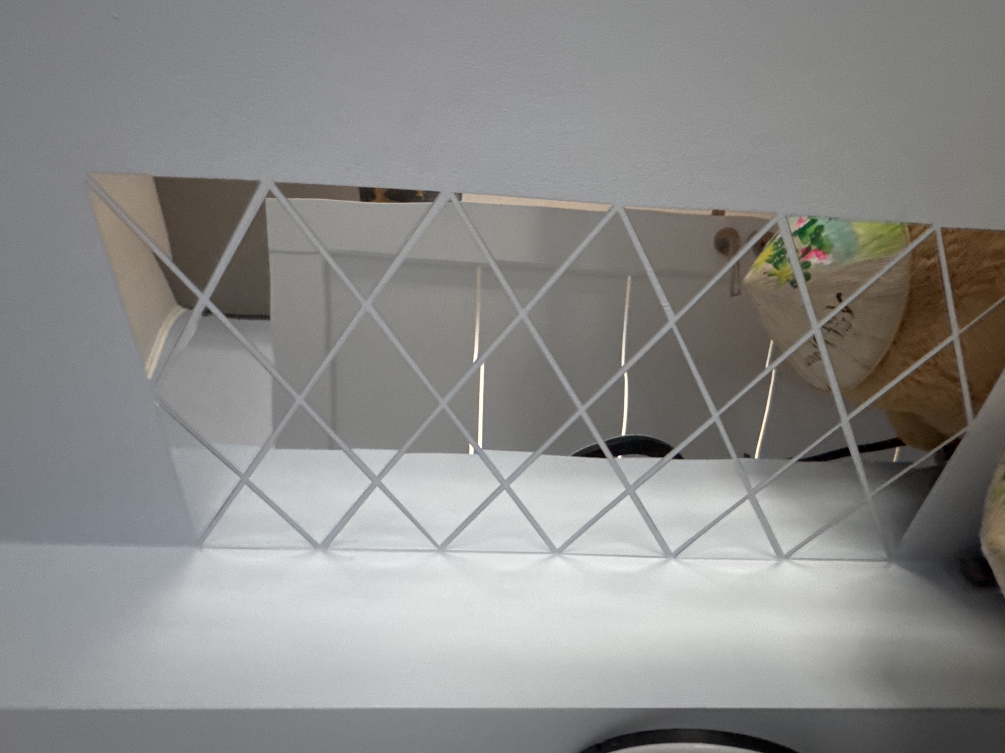 A mirror with a diamond lattice overlay sits on a white shelf. The mirror reflects a white door, part of a black chair, and a tan stuffed animal with a white and green patterned item on top of it. The shelf is against a light gray wall. The location is Plovdiv, Bulgaria, but there are no objects in the image that can further localize it.