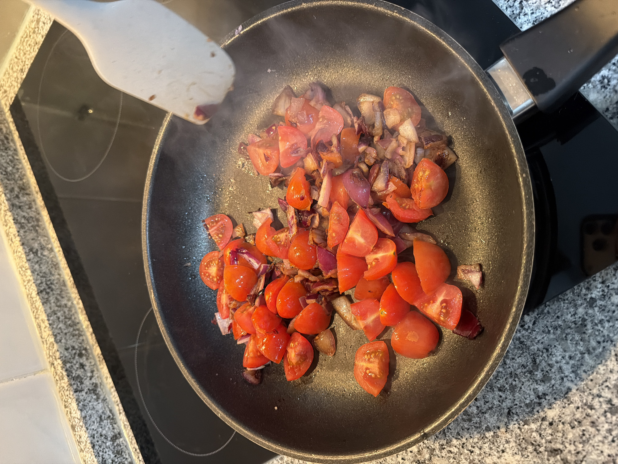 A black frying pan sits on a glass stovetop, containing sauteed diced red onions and halved red tomatoes. A white spatula is positioned above the pan, likely for stirring. Steam rises from the pan, indicating the ingredients are being cooked. The stovetop is part of a kitchen counter with a grey, speckled pattern. A smartphone is visible in the reflection on the stovetop. The scene is likely indoors. There is no verifiable evidence to suggest this image was taken in Vila Real, Portugal.