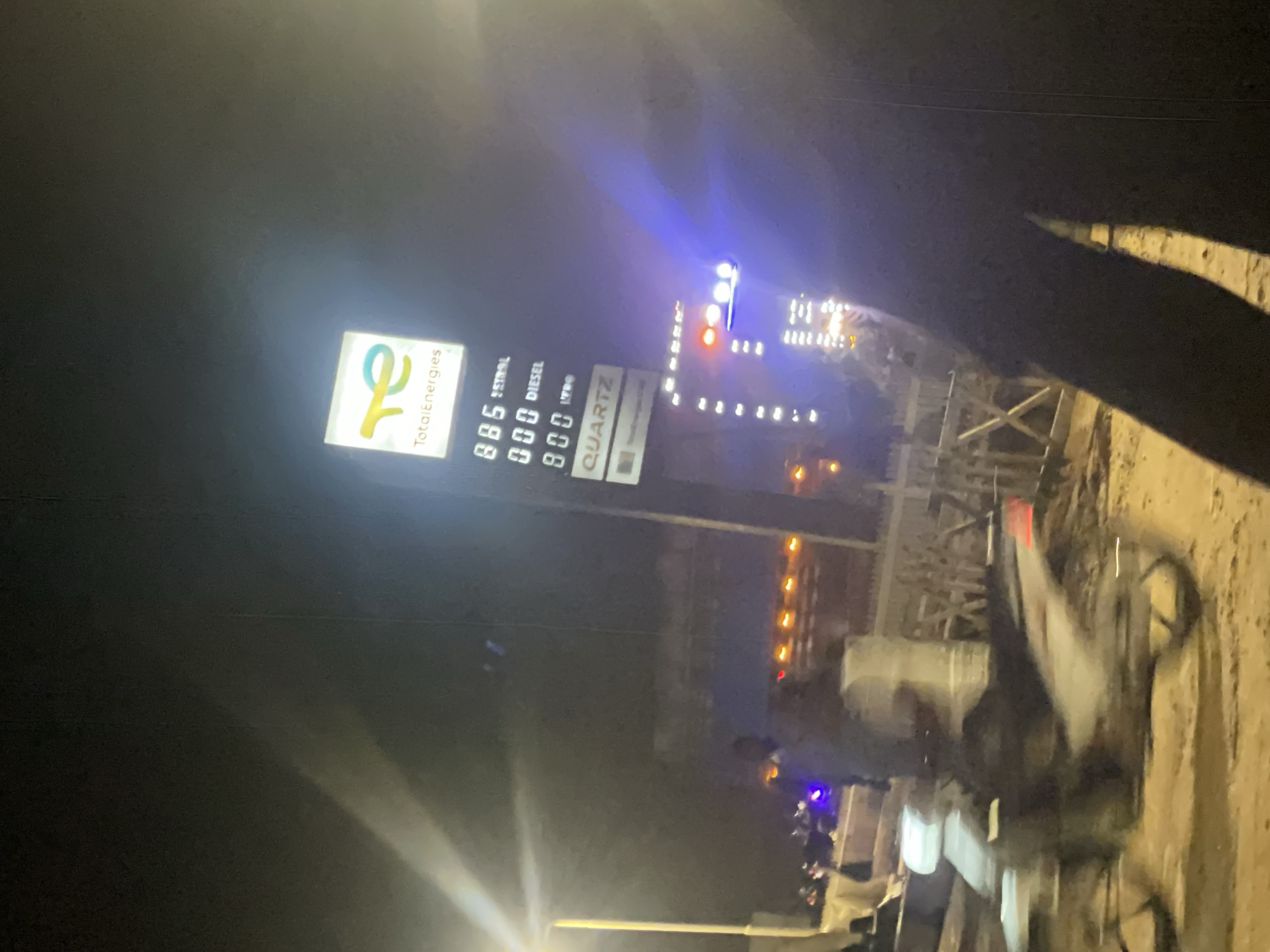 The image, taken at night, captures a TotalEnergies gas station in Lokoja, Nigeria. A lit sign displays "TotalEnergies" and fuel prices: "885 PETROL," "000 DIESEL," and "000 LPG." Below these prices, a "QUARTZ" sign is visible, also from TotalEnergies. In the foreground, a motorcycle is parked, its details blurred due to motion. A person, partially visible, stands near the motorcycle. In the background, other vehicles and a fence are present, illuminated by the gas station's lights. The overall scene suggests activity at the gas station during nighttime hours.
