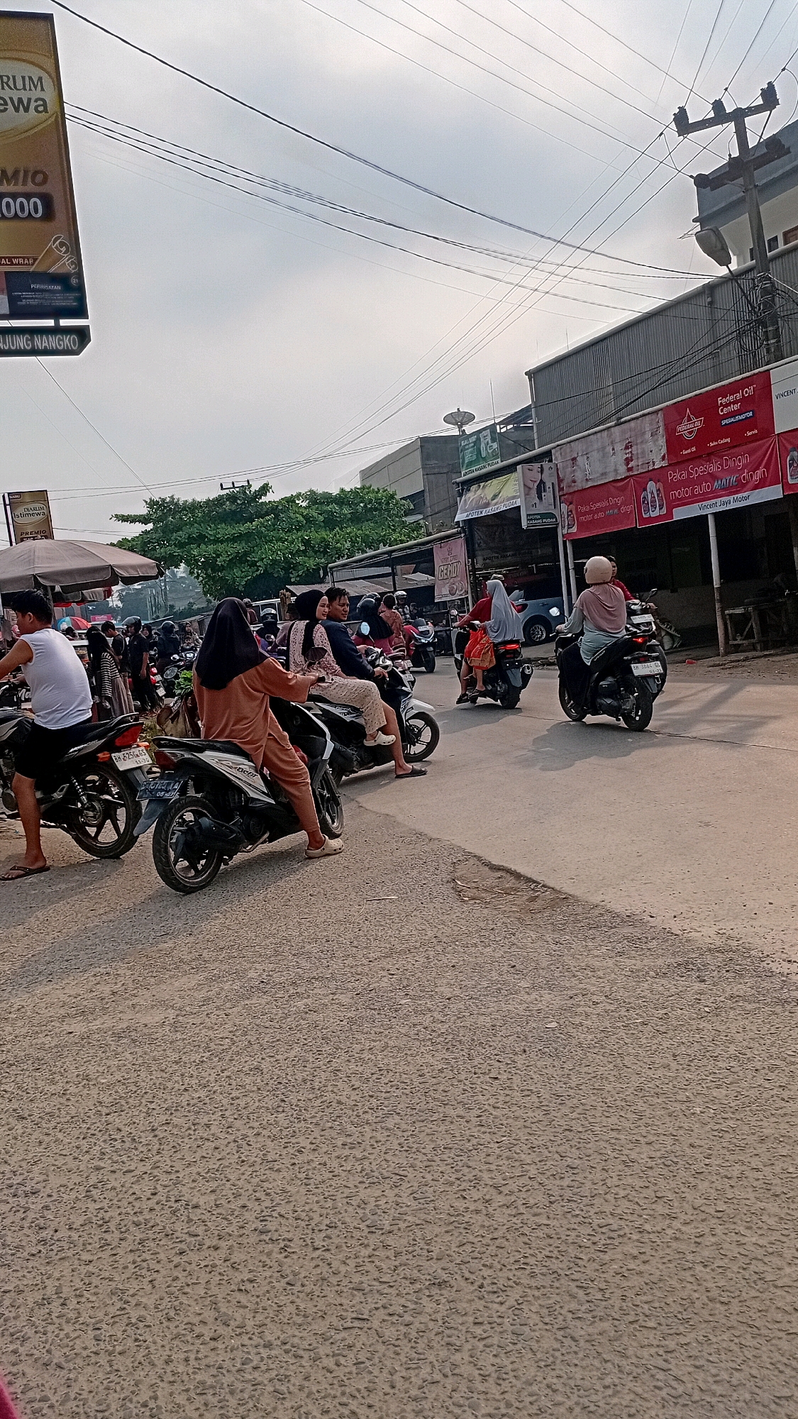 This is a street scene in Kasang Pudak, Indonesia, with several people on motorcycles. The sky is overcast. In the foreground, a person on a motorcycle is seen from behind, wearing a white t-shirt and dark shorts. To their left, another motorcycle is parked. In the middle ground, several people are riding motorcycles, some with passengers. One motorcycle has two women on it, both wearing headscarves and patterned clothing. Another motorcycle has a person wearing a pink helmet and a light-colored shirt. In the background, there are buildings with signs, including one that reads "Federal Oil" and another with red and white lettering. There are also trees and power lines visible. The road surface appears to be asphalt with a gravelly texture.