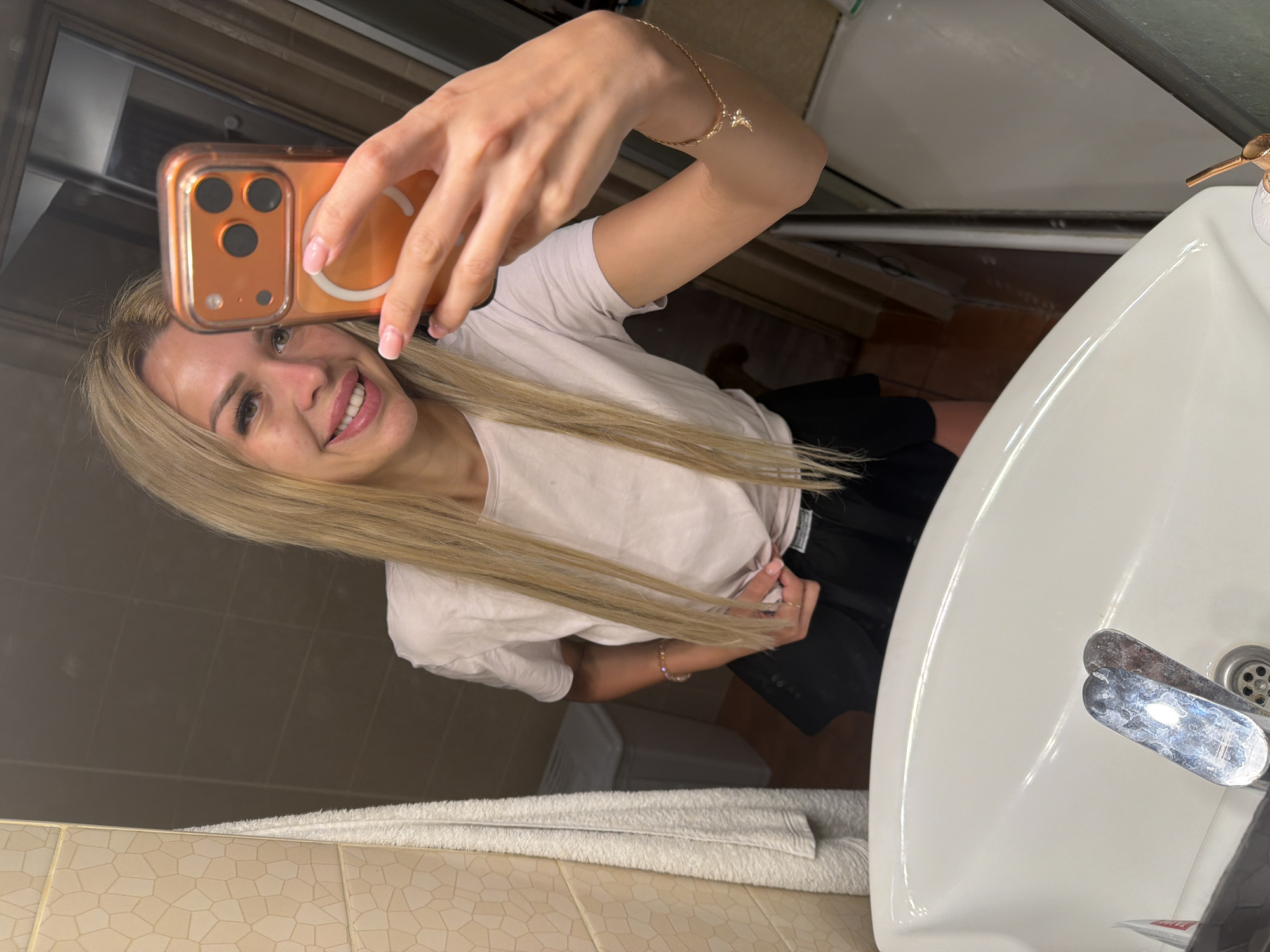 A woman with blonde hair is taking a mirror selfie in a bathroom. She is wearing a light pink t-shirt and black shorts. She is holding an orange iPhone in her right hand, taking the picture. In the background, there is a white sink with chrome faucets, a towel hanging on the wall, and what appears to be a laundry basket. The bathroom walls are tiled with a beige, geometric pattern. The location is a bathroom in Kyiv, Ukraine.
