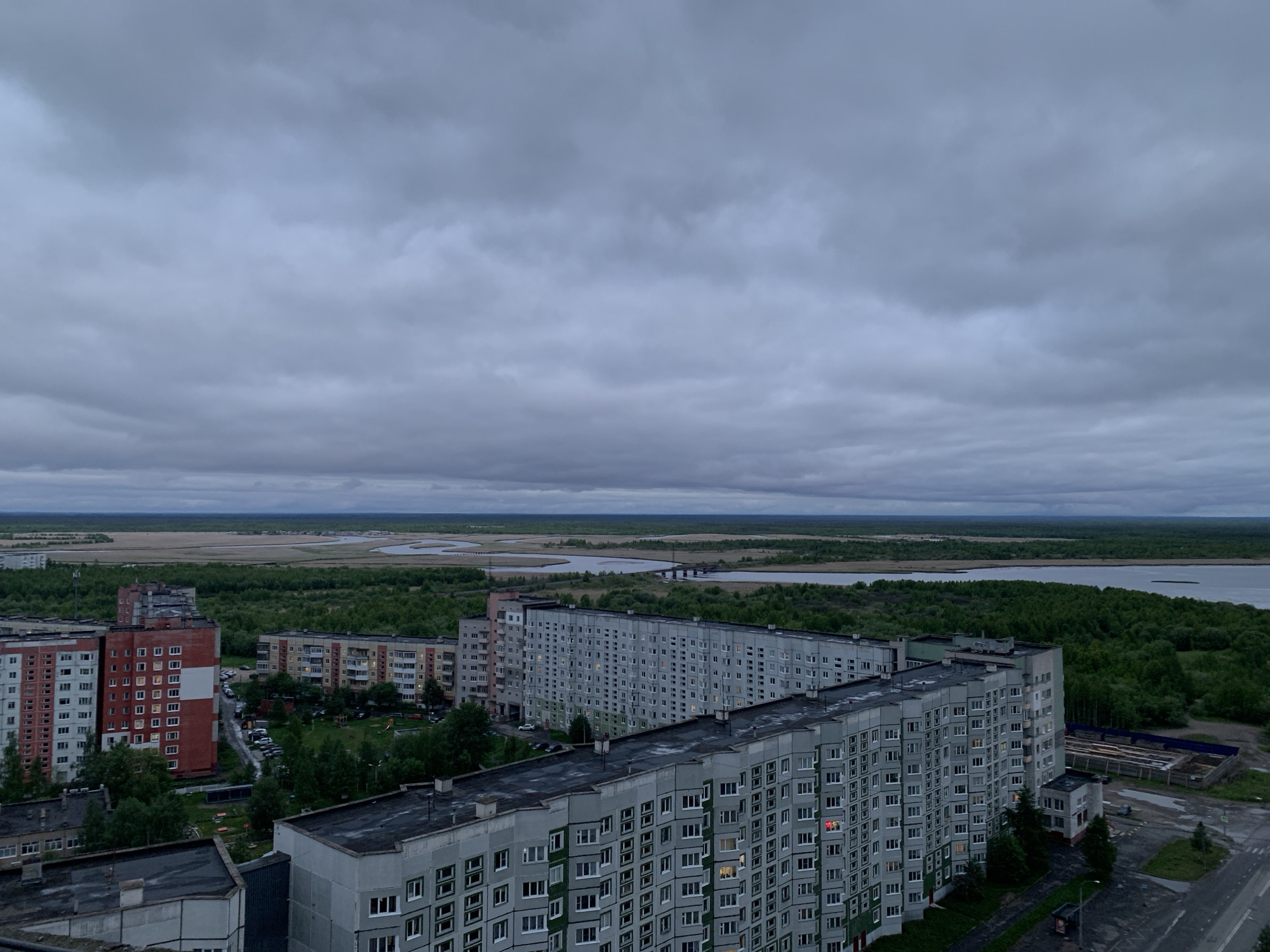 This high-angle, elevated view captures a sprawling urban landscape in Severodvinsk, Russia, under a completely overcast sky. The scene is dominated by numerous multi-story residential apartment buildings, characteristic of post-Soviet architecture, with grey, and some reddish-brown or light-colored facades. Many windows are visible, and a few show interior lights on, suggesting either late evening, early morning, or simply a very dark, cloudy day.
The buildings are arranged in various patterns, forming courtyards with green spaces, sparse trees, and parking lots where several cars are visible. In the mid-ground and background, the urban development transitions into a vast natural landscape. A wide river or estuary meanders through the scene, flanked by extensive, dark green forests. Beyond the forests, lighter, possibly marshy or agricultural land stretches to the horizon. A bridge can be faintly seen spanning the river in the distance.
The sky is a uniform, heavy grey, filled with thick clouds, indicating overcast weather with subdued lighting across the entire scene. No distinct human figures are visible, and there is no discernible text on any structures or signs. The overall impression is one of a functional, densely populated residential area situated on the edge of a significant natural environment, under a muted and somewhat somber sky.