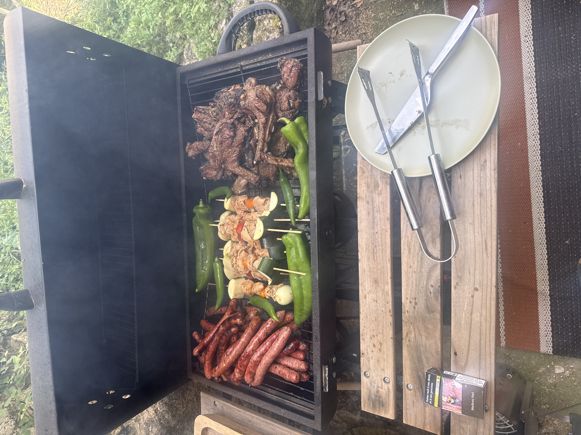 The image shows a barbecue grill outdoors, likely in a residential setting, with food cooking on the grates. There's a pile of cooked meat at the top, skewers of vegetables and meat in the middle, and sausages at the bottom. Green peppers are placed among the sausages and skewers. To the right of the grill is a plate with grilling utensils on top, sitting on a wooden surface. A pack of cigarettes is also visible on the wooden surface. The surrounding environment appears to be a garden or natural outdoor space, with greenery in the background. The location is identified as La Roque-d'Anthéron, France.