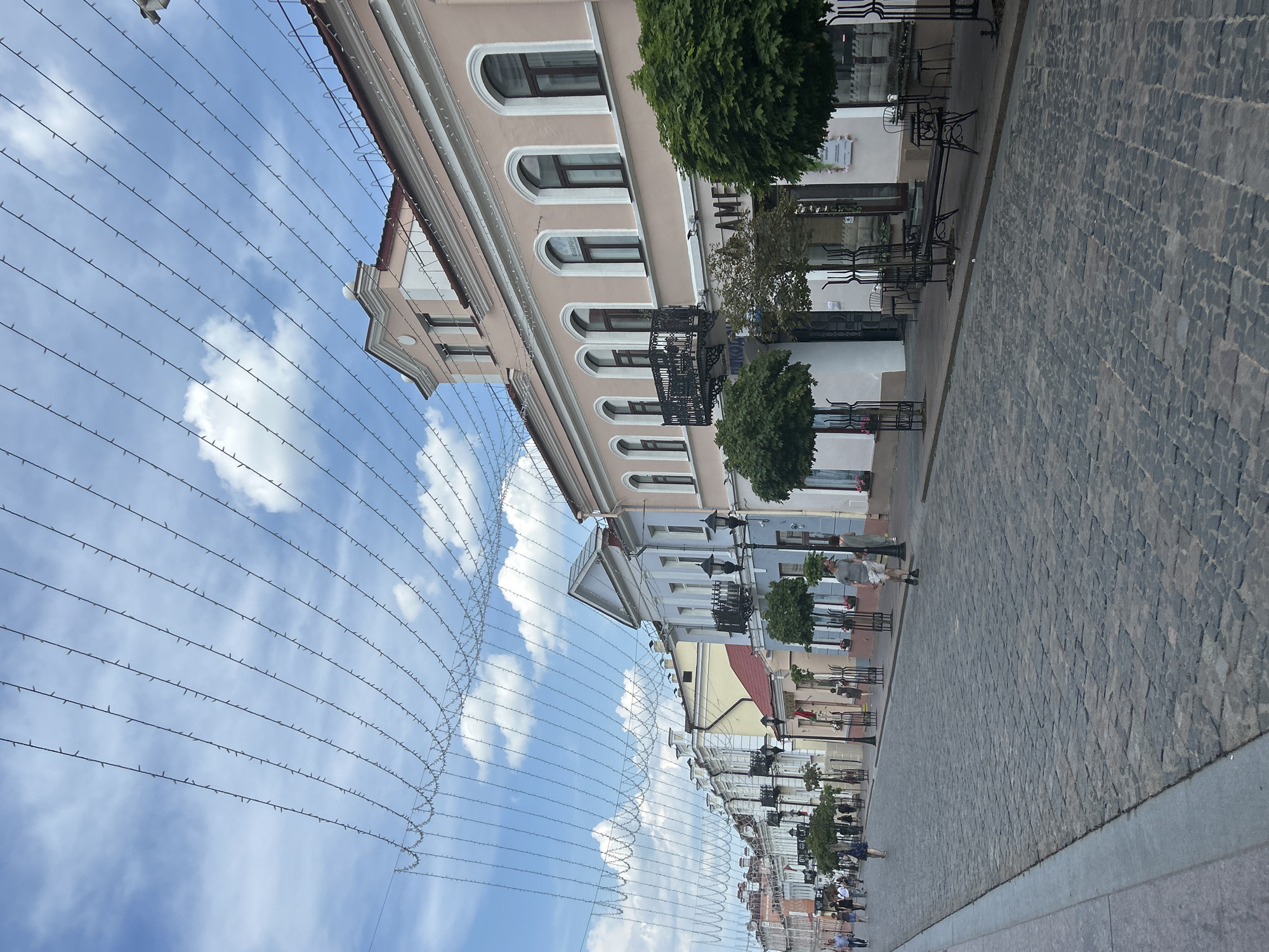 An eye-level, wide shot captures a cobblestone street lined with buildings and strung with lights in an unknown city. The street is paved with dark gray cobblestones, and a gray concrete sidewalk runs along the left side. A few people walk on the sidewalk and street. In the background, a row of historic buildings with pastel facades and pitched roofs stretches along the street. String lights are suspended across the street, creating a decorative canopy. The sky is blue with white, fluffy clouds. Trees are planted along the sidewalk in front of the buildings. The overall scene suggests a picturesque street in a European city.