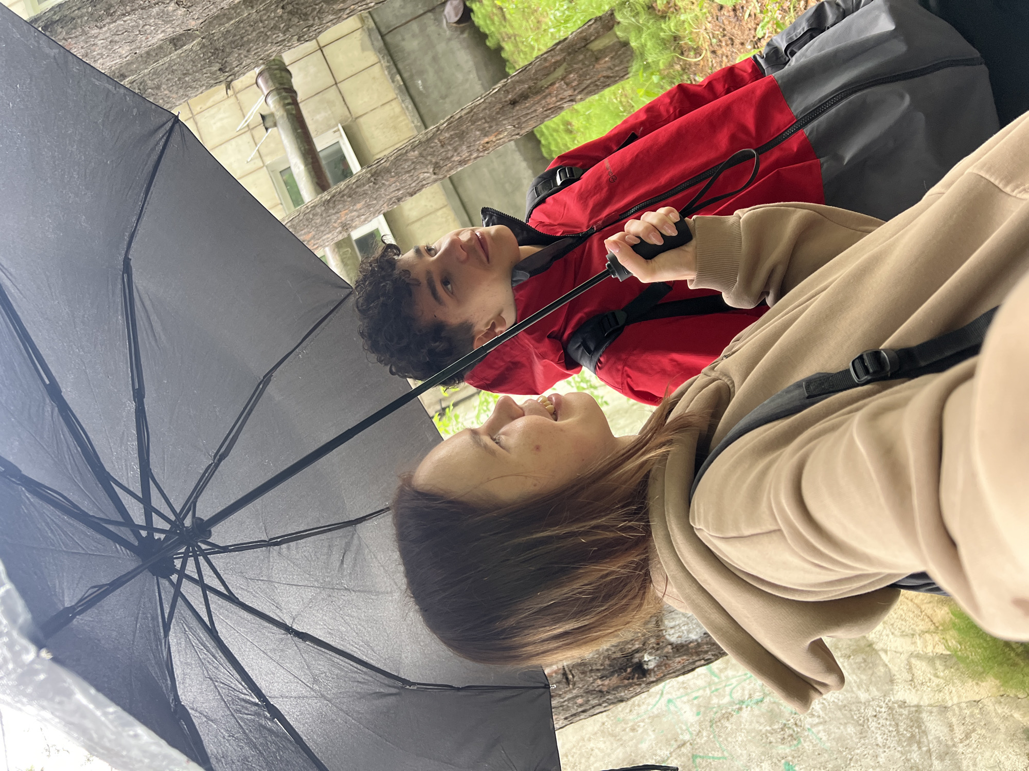 The image captures two young individuals sharing a large black umbrella outdoors in Chaikovsky, Russia, likely during rainy weather as the umbrella appears wet. The perspective is from beneath the umbrella, looking up towards the subjects.
On the left, a young man with dark, curly hair and light brown eyes looks towards the right with a gentle smile. He is wearing a red and dark grey two-toned jacket, possibly a waterproof or windbreaker style, with a visible black zipper and shoulder strap from a backpack.
On the right, a young woman with light brown hair is smiling, looking towards the man. She wears a beige hooded sweatshirt and holds the umbrella's handle with her right hand, which has neatly manicured nails. A dark strap, likely from a backpack, is visible over her shoulder.
The setting includes natural elements like tree trunks with textured bark and green foliage, alongside urban features such as a light-colored building with windows and a drainpipe in the background. The ground appears to be a mix of grass and possibly concrete or paved areas. The expressions of both individuals suggest a pleasant and amicable interaction. There is no discernible text visible in the image.