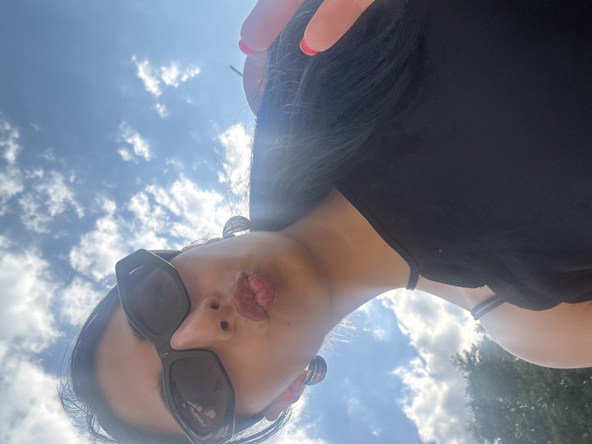 A young woman, wearing dark sunglasses and a black top, puckers her lips and holds up two fingers in a peace sign. She is outdoors, with a bright blue sky and scattered clouds in the background. Trees are visible in the lower left of the frame. The image appears to be a selfie, taken from a low angle looking up at the woman. There are no discernible Plovdiv, Bulgaria landmarks or features visible in the image.