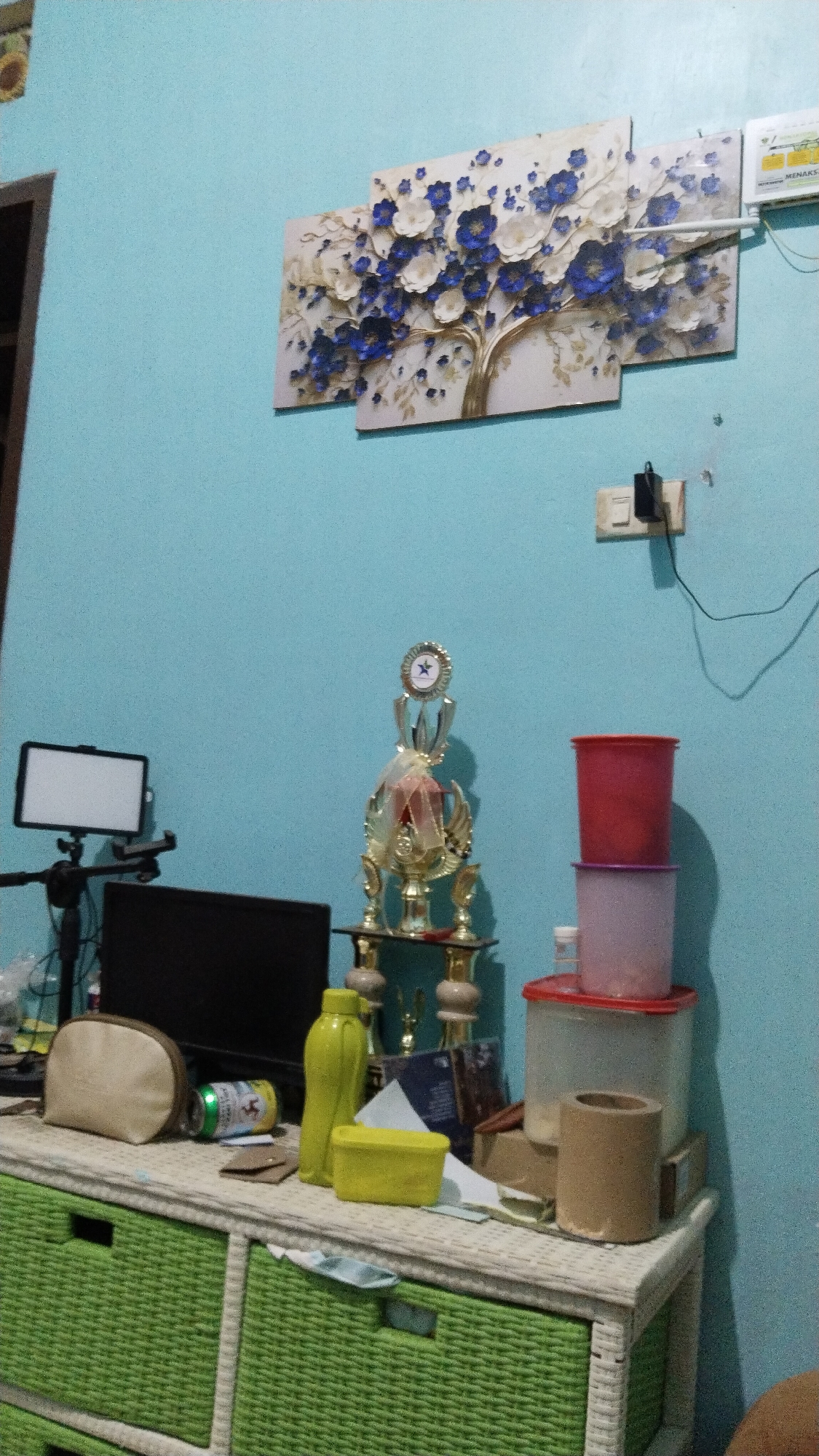 The image depicts an indoor scene, likely a room, with a light blue wall as the backdrop. On the wall, a three-panel artwork of a tree with blue and white flowers is mounted. To the right of the artwork, an electrical outlet with a plug is visible.
In the foreground, a white woven dresser with green drawers occupies the lower portion of the image. On top of the dresser, several objects are arranged. From left to right, there is a light-colored makeup bag, a silver and green can, a yellow water bottle, a yellow rectangular container, and a trophy with gold accents and a red ribbon. Behind these items, a dark computer monitor is partially visible. To the right of the trophy, a stack of three plastic containers of varying sizes and colors (red, purple, and clear) are placed. A cylindrical brown object is also on the dresser.
The scene appears to be a domestic setting. There are no people visible in the image. The lighting suggests an indoor environment, possibly during the day or with artificial lighting.
