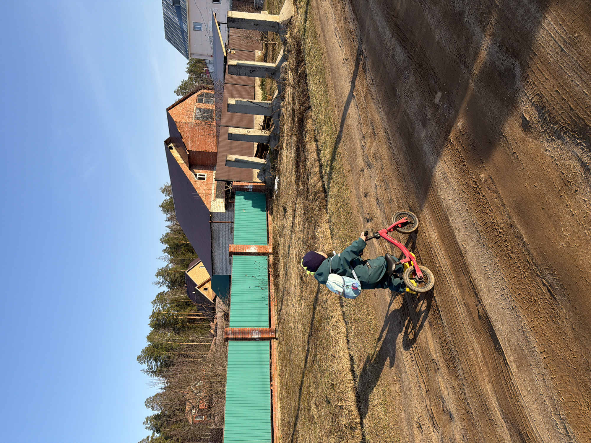 The image depicts a child riding a small red and yellow bicycle on a dirt road in what appears to be a residential area. The child is wearing a dark green jumpsuit with a small, light blue backpack. The road has visible tire tracks. A green metal fence with brick pillars runs along the side of the road. Behind the fence, there are trees and houses, including one with a red brick exterior. The sky is blue. The scene is located in the city of Chaikovsky, Russia.