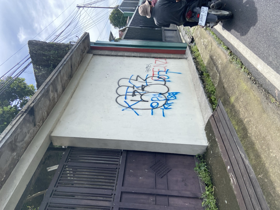 The image shows a white concrete wall with graffiti on it. The graffiti is in blue and black paint and appears to be gang-related. The wall is located on a residential street in Rancabali, Indonesia, at the coordinates -7.119, 107.433. There is a brown wooden gate to the left of the wall and a motorcycle parked on the street to the right of the wall. A man in a blue shirt and khaki pants is sitting on the motorcycle, with his back to the camera. The street is paved with asphalt and has a white stripe painted down the middle. The sidewalk next to the wall is made of concrete and has a brown wooden bench on it. The environment is suburban, with houses and fences in the background. The sky is cloudy.