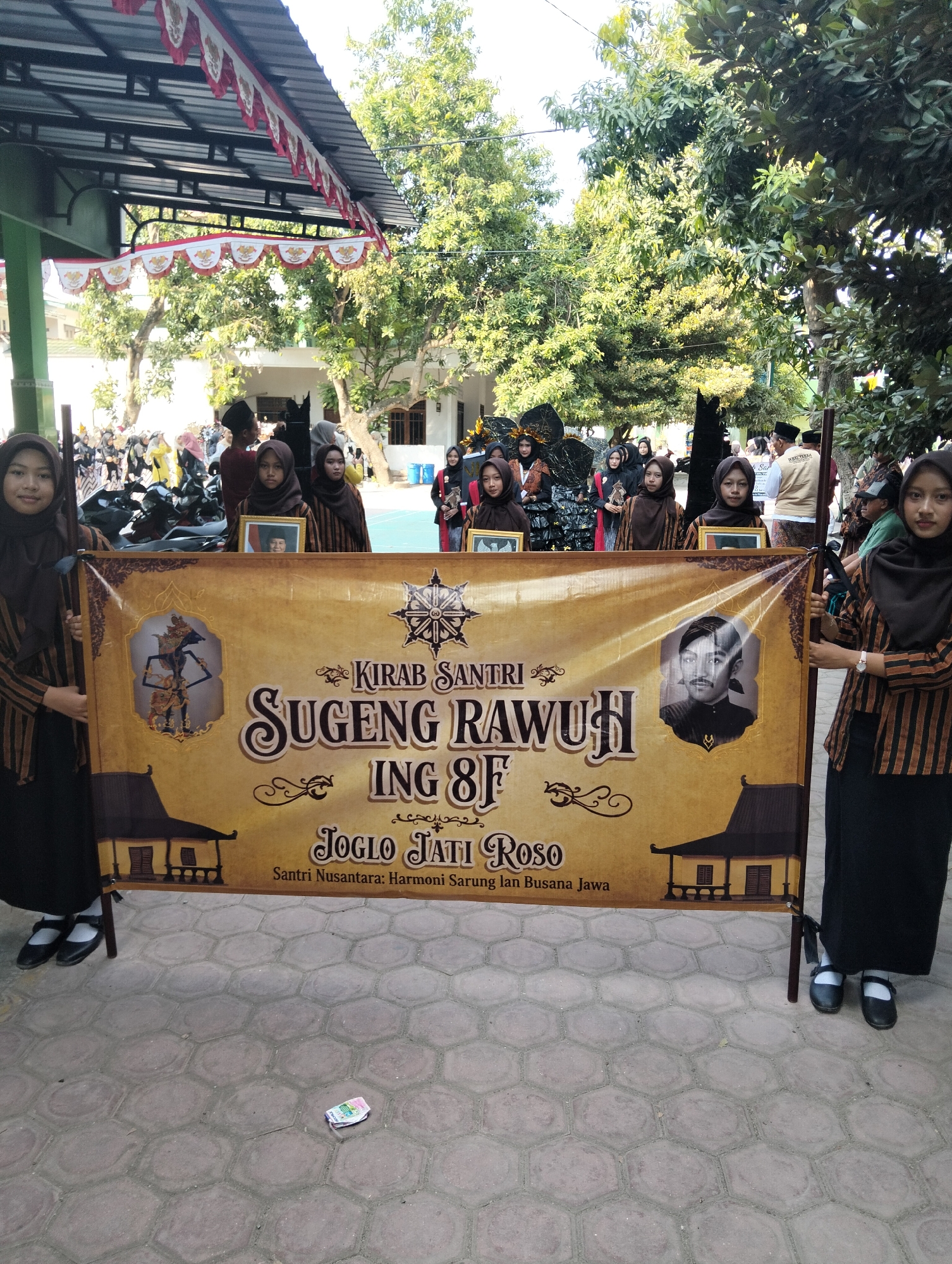 In Kranji, Indonesia, four young women in matching brown and black striped uniforms and headscarves stand on a paved courtyard holding a large, ornate banner. The banner is golden yellow with dark brown decorative elements and text. It features a stylized wheel emblem, a puppet-like figure in a frame on the left, a portrait of a man with a mustache in a frame on the right, and an illustration of a traditional Indonesian house. The text on the banner reads: "KIRAB SANTRI SUGENG RAWUH ING 8F JOGLO JATI ROSO Santri Nusantara: Harmoni Sarung lan Busana Jawa". In the background, a group of people, some in traditional attire and others in casual clothing, are visible, along with motorcycles parked behind them. Overhead, a roofed structure is adorned with red and white banners. The ground is covered with hexagonal paving stones, and a small wad of cash is lying on the pavement.