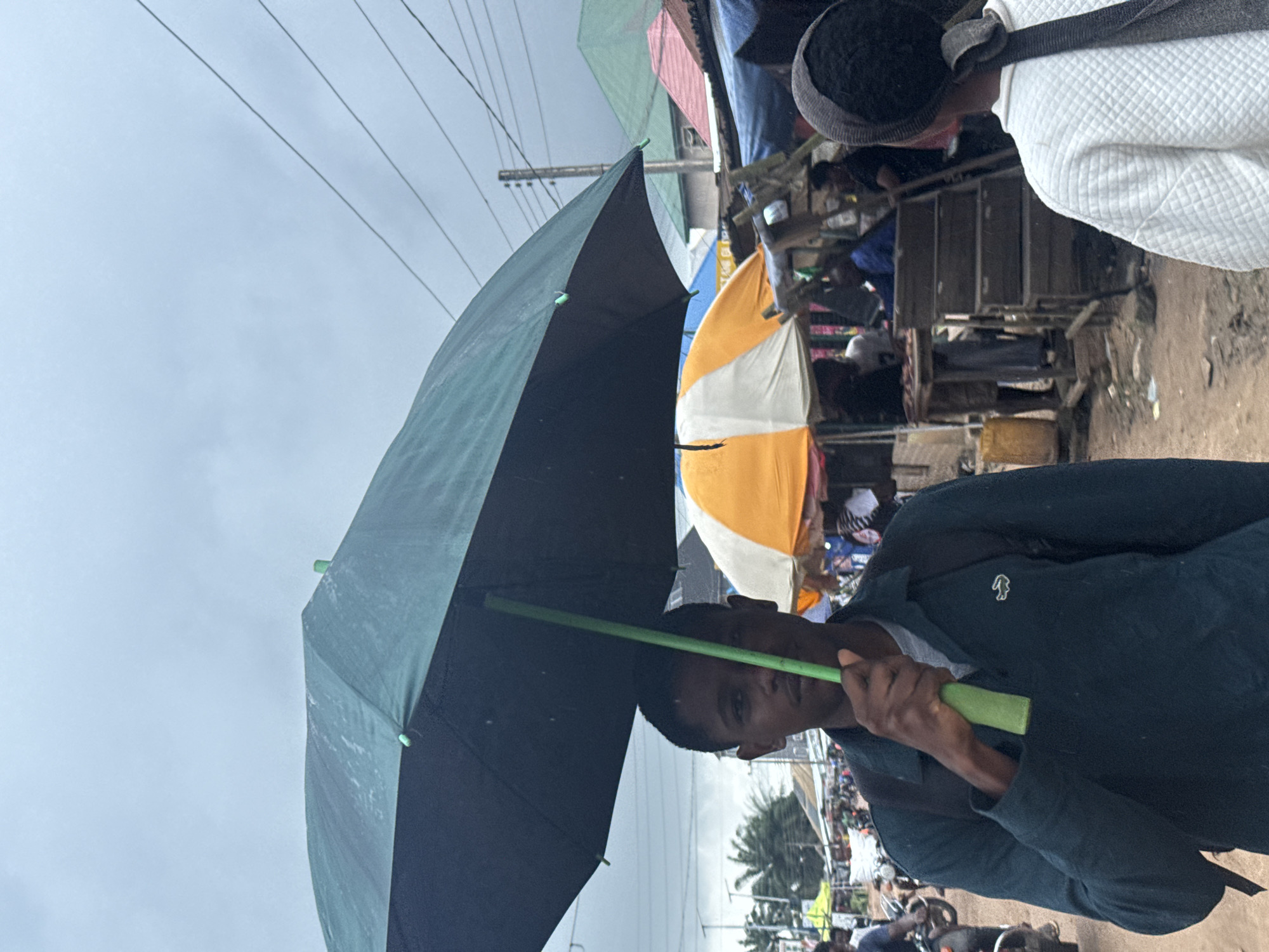 In Ihiagwa, Nigeria, under a cloudy, overcast sky, a young man stands holding a large, dark green umbrella over his head. He is wearing a dark long-sleeved shirt with a small white alligator logo on the left chest and a grey t-shirt underneath. A black backpack is visible on his back. The umbrella has a bright green handle and metal accents.
The background depicts a bustling outdoor market scene. Several stalls are visible, covered by various umbrellas and awnings. To the right of the main subject, an orange and white umbrella provides cover for a stall. Further back, there are more market stalls with people tending to them or browsing. Structures with corrugated metal roofs in various colors, including green and red, are also visible. In the middle ground, several motorcycles are parked, suggesting the presence of transportation in the area. The ground appears to be dirt or gravel, typical of an unpaved market area. The overall atmosphere suggests a community market operating, possibly during or after a rainfall, given the presence of umbrellas and the cloudy sky.