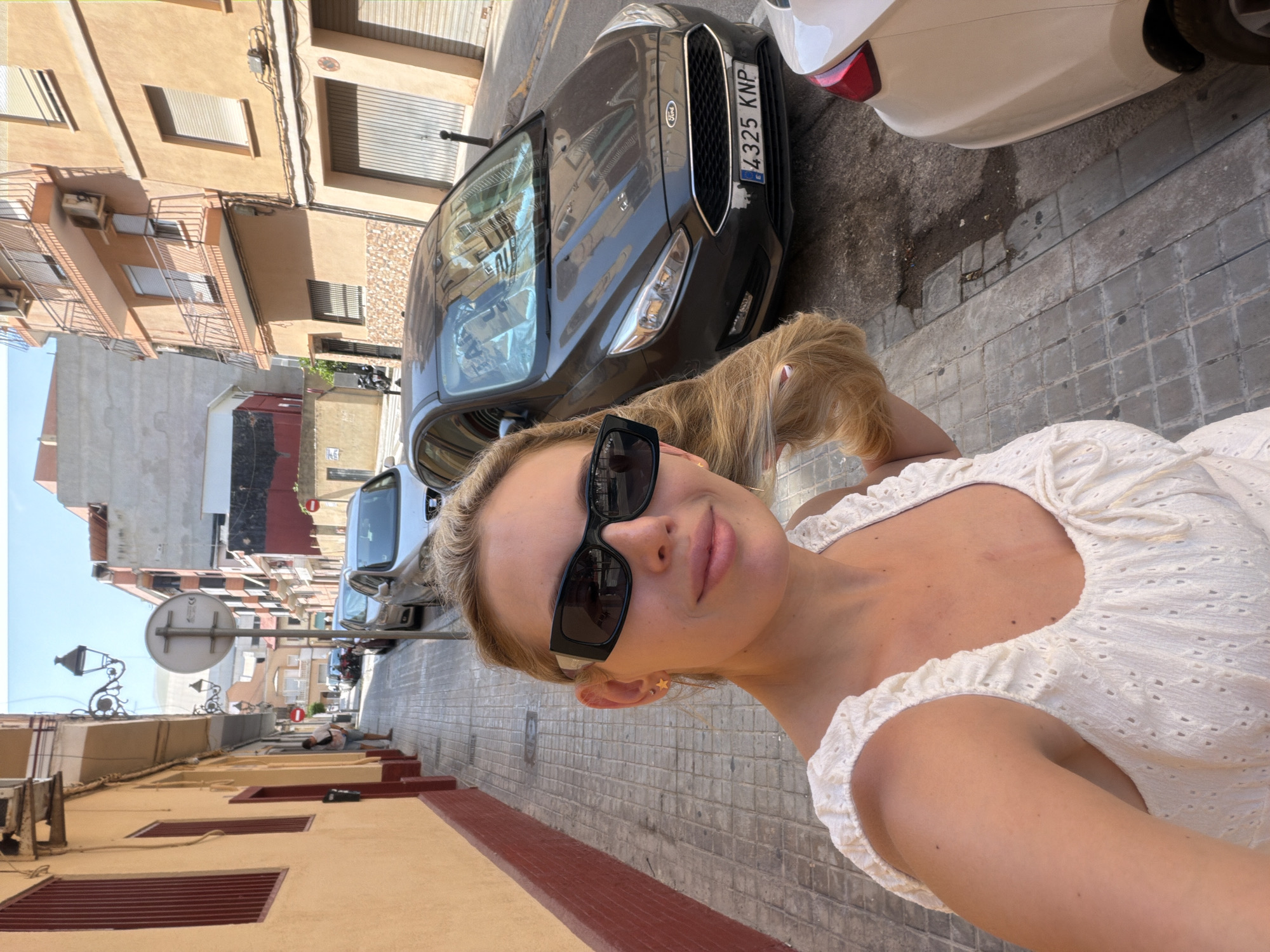 The media file is a selfie taken by a young woman with blonde hair, wearing a white eyelet dress and sunglasses. She is looking at the camera and smiling. The background shows a narrow street in a European city, with buildings on either side and parked cars. One car, a dark Ford, has a license plate that reads "4325 KNP". The street is paved with gray cobblestones, and there are streetlights and a traffic sign visible. The buildings are a mix of light brown and beige, with balconies and windows. The overall impression is a sunny day in a Mediterranean town. The woman's attire and the outdoor setting suggest a casual, pleasant day. The woman's expression conveys happiness and confidence. The lighting suggests it is daytime, likely mid-morning or afternoon, due to the bright sunlight casting shadows. The architecture and street scene are consistent with a location in Spain, and specifically Valencia, as indicated by the context.