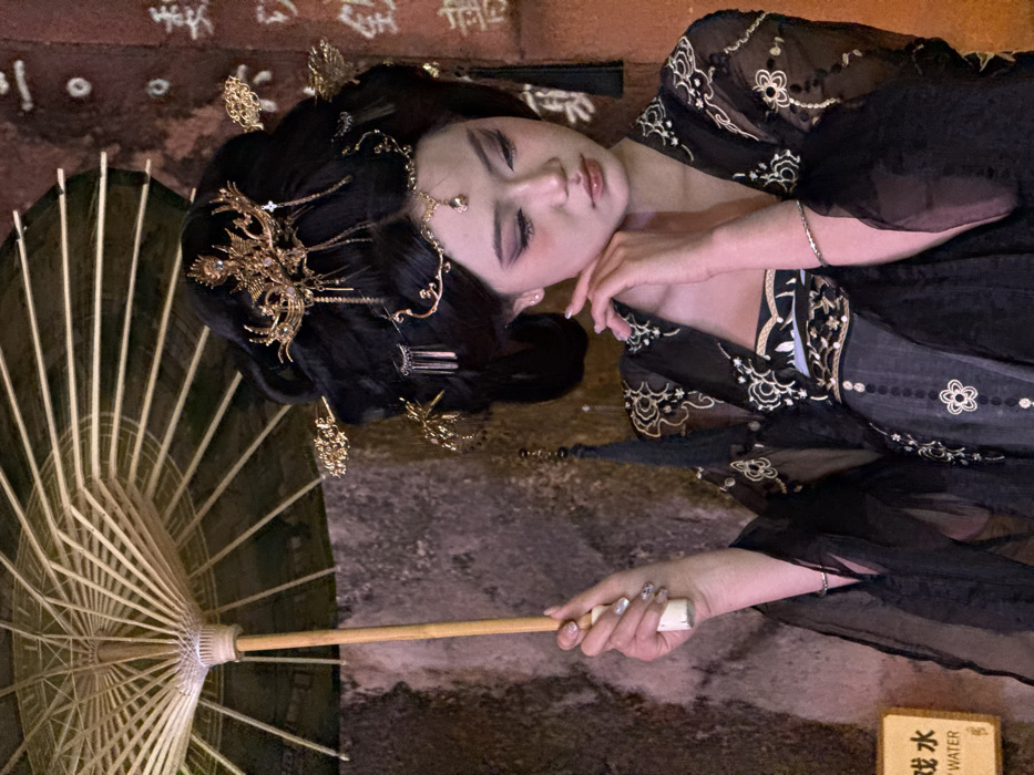 The image displays a young East Asian female, positioned slightly off-center to the right, in a dimly lit, traditional setting within Yuzhong District, China. She wears an elaborate dark-colored traditional Chinese-style outfit, featuring a sheer black outer garment with intricate gold and white embroidery, layered over darker undergarments. Her dark hair is styled in a voluminous updo, richly adorned with multiple golden hairpins, a prominent golden crown-like ornament, and a forehead jewelry piece with a central gem. Her makeup includes defined eyes and blush. Her right hand, with manicured nails, holds the wooden handle of an open, dark-canopied paper parasol. Her left hand is raised, with fingertips resting near her chin, adopting a contemplative pose with her gaze directed downwards. The background consists of textured walls: on the left, a darker, possibly stone wall with a yellow rectangular sign displaying "戏水" (xì shuǐ) and "Y WATER"; on the right, a reddish-brown textured wall with vertically written, partially visible Chinese characters, including "楼" (lóu) and "街" (jiē). The lighting is localized, highlighting the subject against the darker environment.
