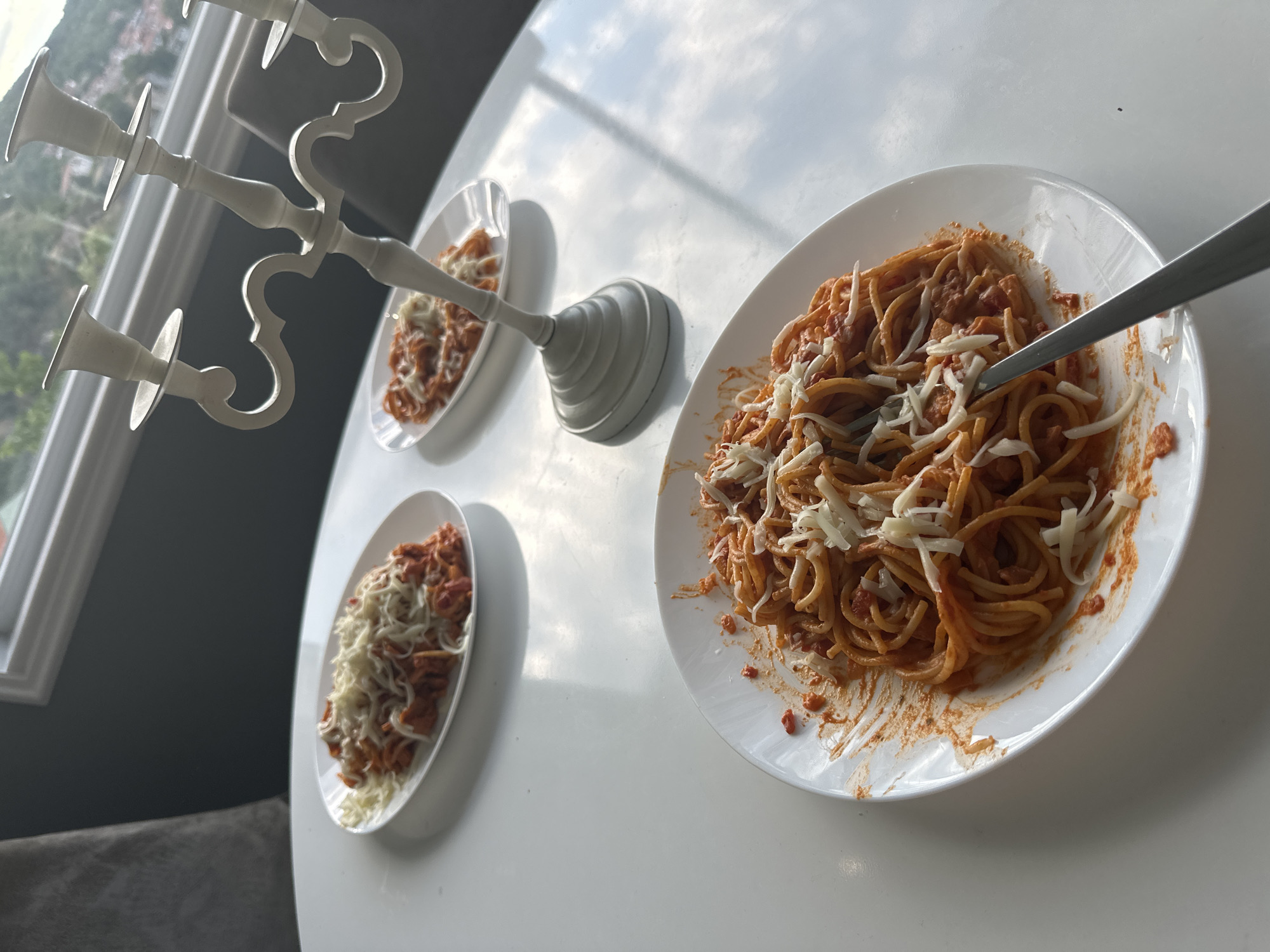 The image shows three plates of spaghetti with tomato sauce and shredded cheese arranged on a white table. A white candelabra is positioned to the left of the plates. One plate of spaghetti is prominently featured in the foreground on the right, with a fork in it. Two smaller plates of spaghetti are positioned to the left, behind the candelabra.
The setting appears to be indoors, with a window in the background showing a glimpse of trees and buildings, suggesting a daytime scene. The lighting suggests it's likely late morning or early afternoon, given the bright sunlight streaming in and casting shadows. The overall mood is relaxed and informal, perhaps indicating a home-cooked meal. There is no visible text in the image.