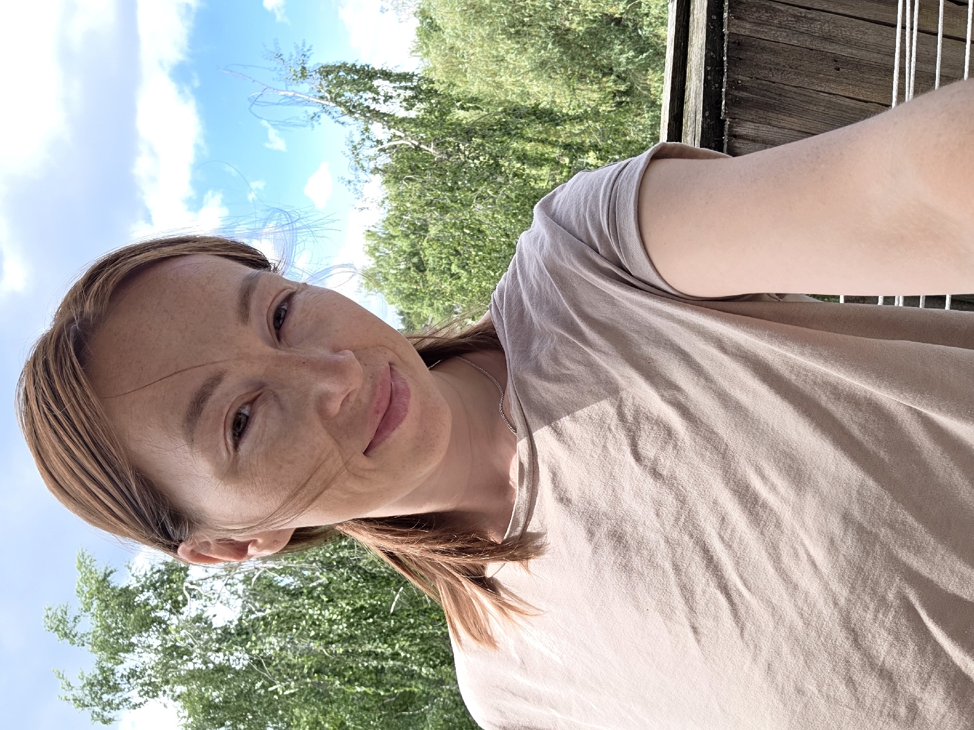 A woman with reddish-brown hair tied in a ponytail is taking a selfie outdoors. She is wearing a light brown t-shirt and a silver necklace. Her face is turned slightly towards the camera, and she is smiling. Behind her, there are lush green trees and a blue sky with white clouds. The scene appears to be on a balcony or a wooden deck, indicated by the wooden railing in the foreground. Based on the context provided, this image was taken in Chaikovsky, Russia.