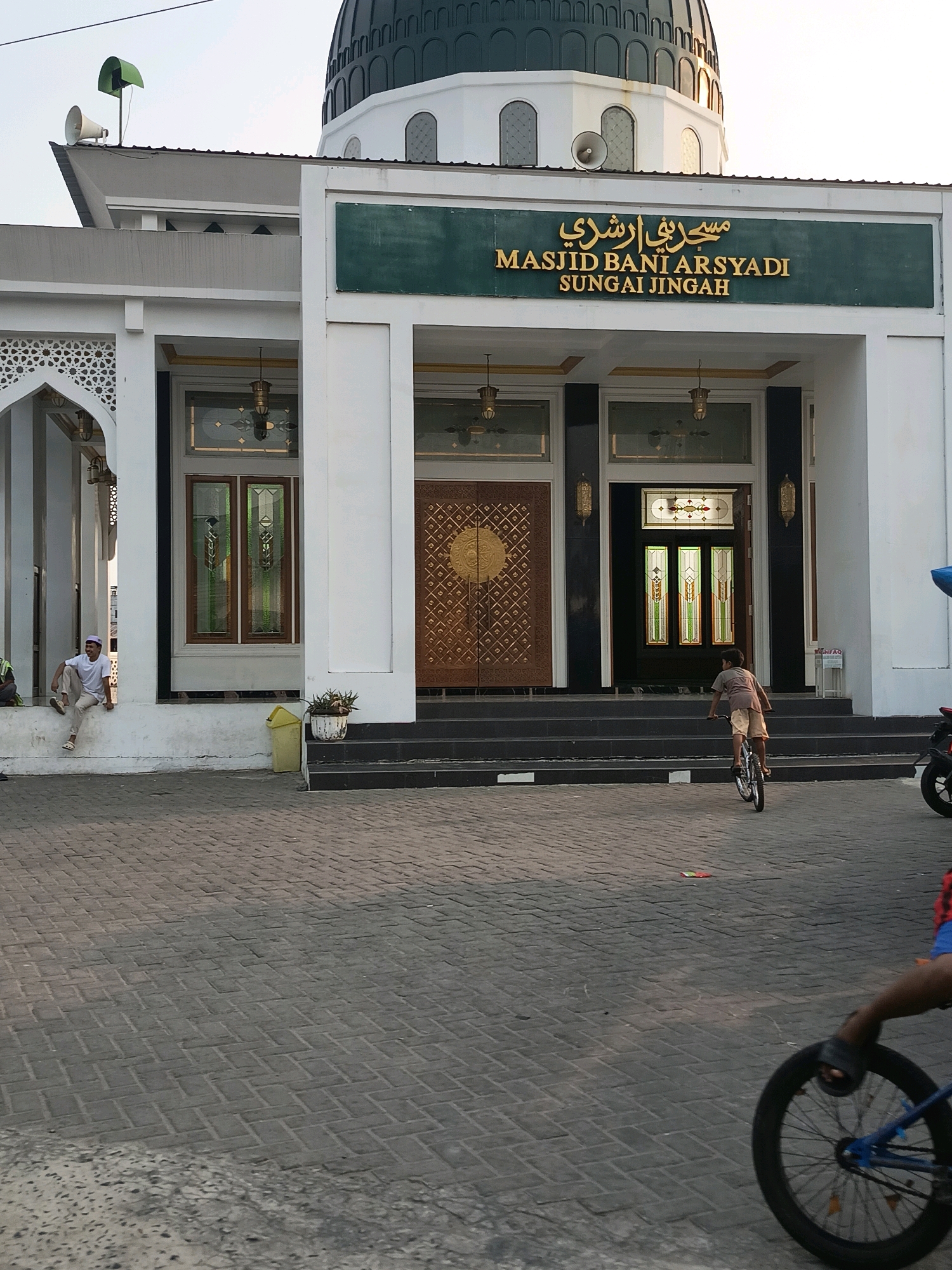The image depicts the entrance of the Masjid Bani Arsyadi in Sungai Jingah, Banjarmasin, Indonesia. The mosque features a prominent green dome and a white facade with intricate architectural details, including arched windows and decorative carvings. The entrance is grand, with large wooden doors adorned with geometric patterns and flanked by ornate stained-glass windows.
In the foreground, a young boy is seen riding a bicycle on a paved courtyard. He is wearing a t-shirt and shorts. To the left, a young man is sitting on a ledge, wearing a white t-shirt and pants with a white cap. He appears relaxed, looking towards the left side of the frame. A yellow trash can is visible near the steps leading up to the mosque. The courtyard is paved with interlocking brick pavers.
The lighting suggests it is daytime, possibly late afternoon, with the sun casting shadows across the scene. The overall atmosphere is calm and serene, with a touch of everyday life unfolding around the religious building.