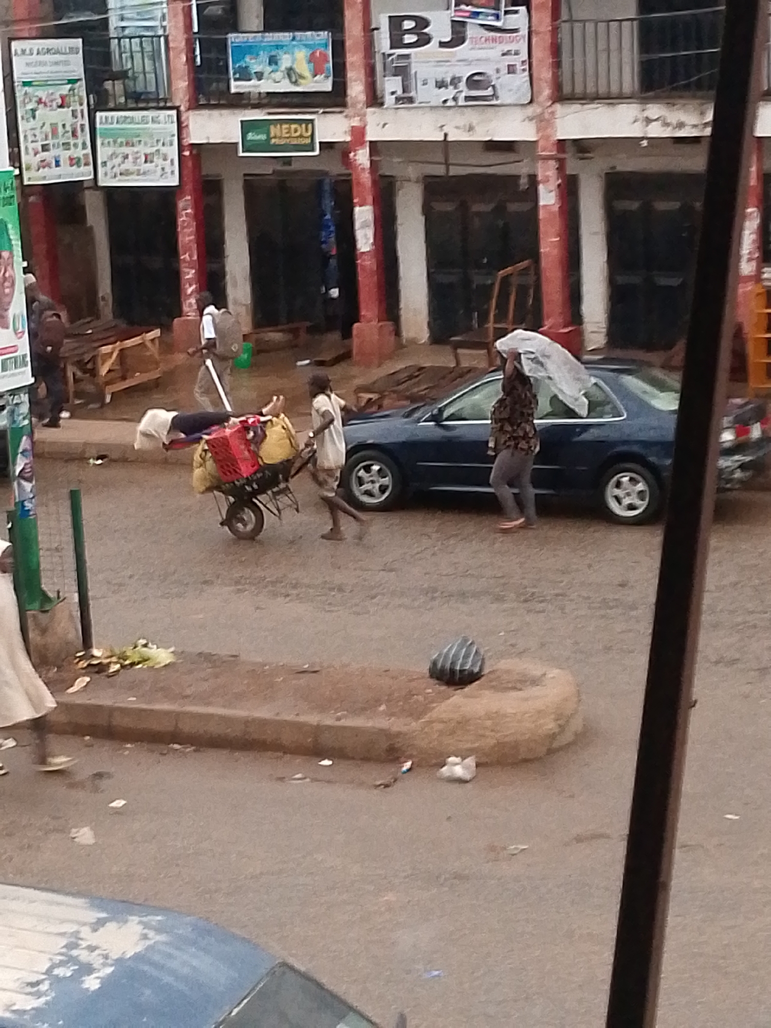 The scene takes place on a street in Bukuru, Nigeria, with a two-story building in the background, displaying signs such as "A.M.B. AGROALLIED" and "NEDU PROVISION". A man is pushing a wheelbarrow laden with goods, including a red crate, yellow bags, and what appears to be a person lying on top, covered with a white sheet. Next to the wheelbarrow and parked car, a woman is holding a white cloth or plastic sheet over her head, likely for protection from the sun or rain. The street is paved but appears worn. Other individuals are present in the background, near some wooden structures. In the foreground, there's a section of curb with some litter and a striped bag. The overall lighting suggests daylight, and the scene captures everyday street activity in an urban environment.