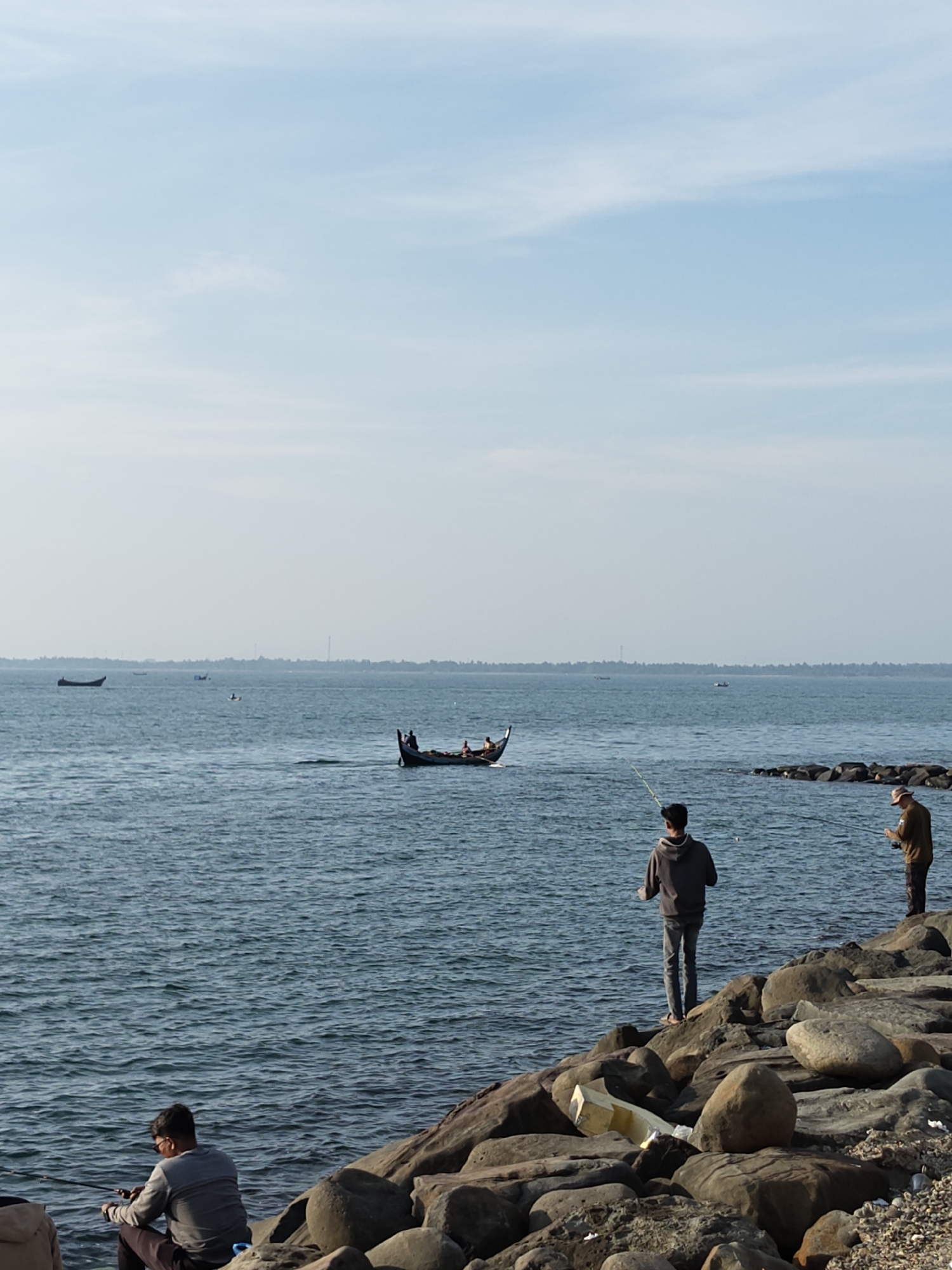This vertical-oriented image presents a serene coastal scene in Lhokseumawe, Indonesia, under a bright, light blue sky with sparse, wispy clouds, suggesting a clear day.
The foreground is characterized by a rocky shoreline composed of large, dark, irregularly shaped boulders. Three men are engaged in fishing from these rocks. On the right, one man in a dark hoodie and jeans stands, holding a fishing rod pointed towards the water. Beside him, further to the right, another man wearing a hat and a light brown shirt also stands, fishing. In the lower-left corner, a third man in a grey long-sleeve shirt and sunglasses sits on a rock, intently holding his fishing rod.
The mid-ground features a calm, deep blue sea with gentle ripples. A traditional-style boat with a dark hull and pointed ends, carrying several people, is visible moving across the water from left to right. Several other smaller boats are scattered further in the distance, closer to the horizon. A faint, distant coastline with a subtle silhouette of land and structures stretches across the background, indicating the presence of land on the far side of the water.
The scene conveys a peaceful atmosphere, highlighting both recreational fishing and local maritime activity. No discernible text is visible within the image.