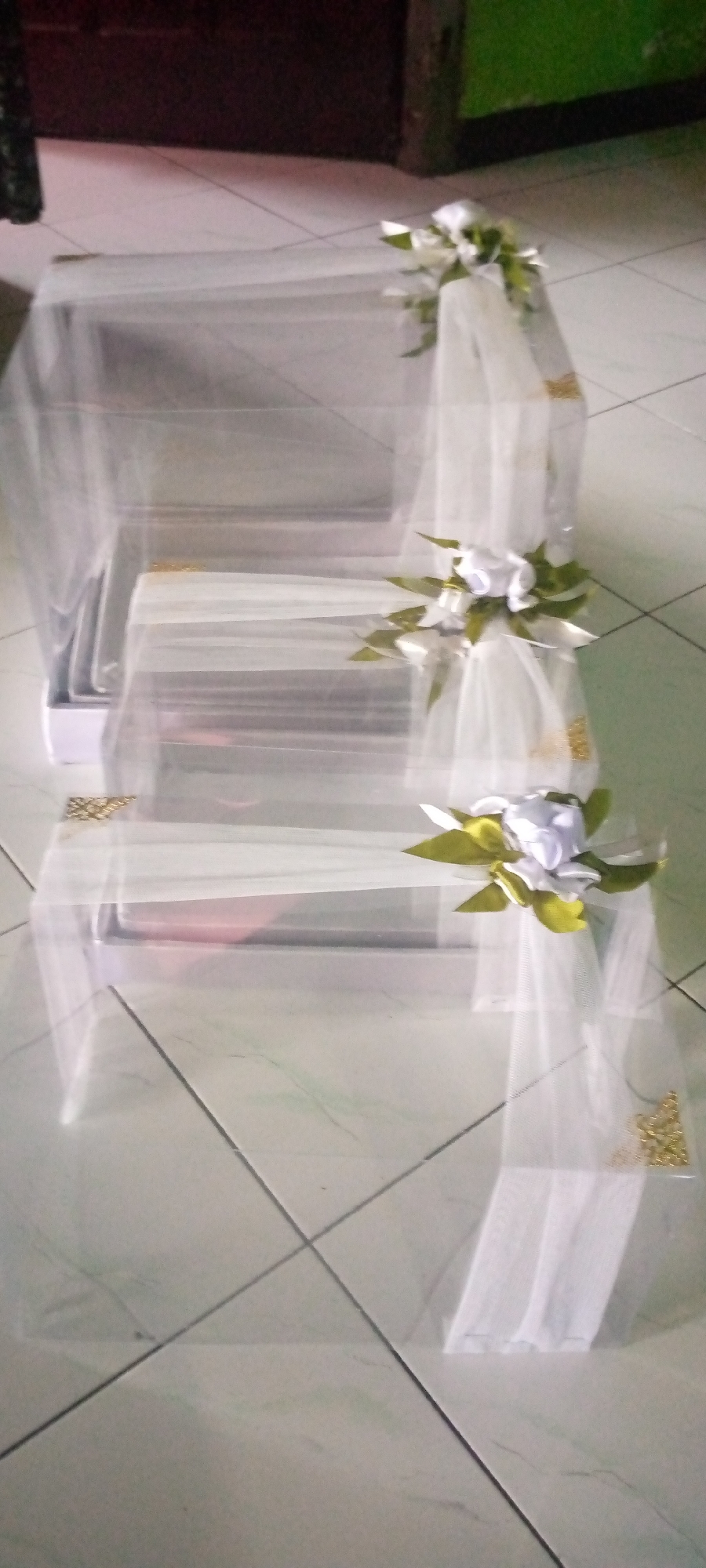 The image shows three rectangular boxes stacked vertically, draped in white sheer fabric and decorated with white artificial flowers and green leaves. The boxes appear to be gift boxes. The bottom box has a hint of pink visible inside. Gold decorative accents are present on the corners of the boxes and along the fabric. The boxes are arranged on a light-colored tiled floor, with dark brown and green walls visible in the background. The scene is likely an indoor setting, possibly a room in a home in Sumberwono, Indonesia, given the tiled flooring and wall colors common in Indonesian homes. There are no people visible in the image.