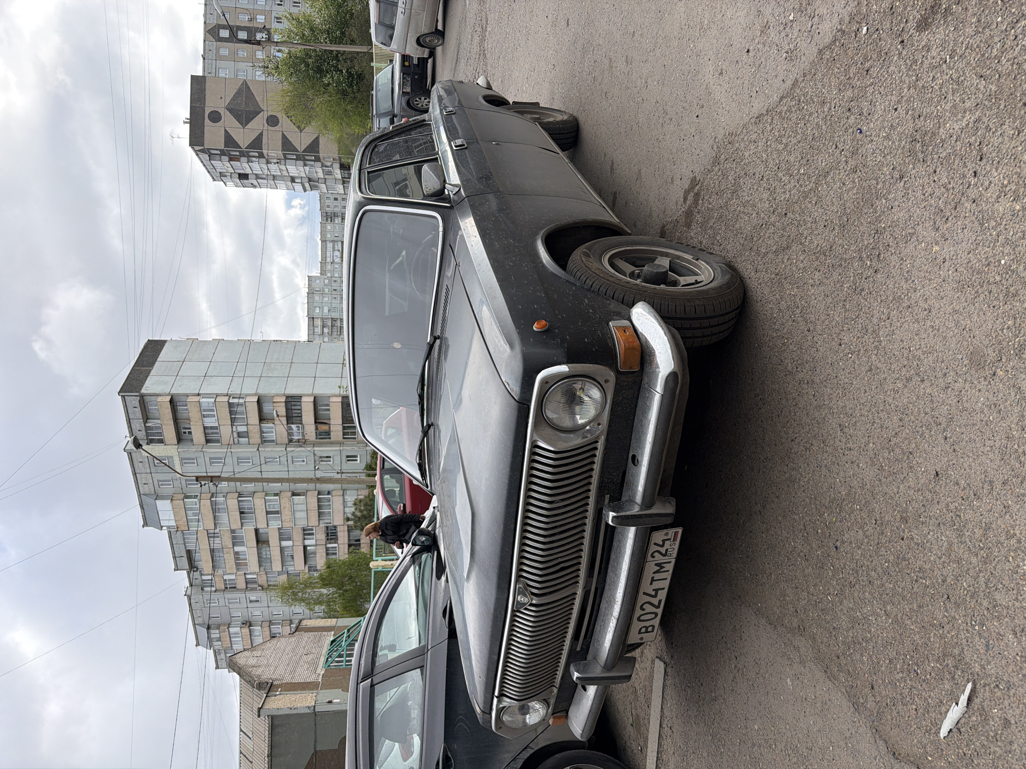 The image shows a black vintage car parked on an asphalt street, with multi-story apartment buildings in the background. The car has a chrome grill and bumper, and a Russian license plate reading "B 024 TM 24 RUS". Another vehicle is parked behind it. The apartment buildings are typical Soviet-era structures. A person stands beside a car in the background. Power lines stretch across the sky. The scene is in Krasnoyarsk, Russia, indicated by the license plate code "24".