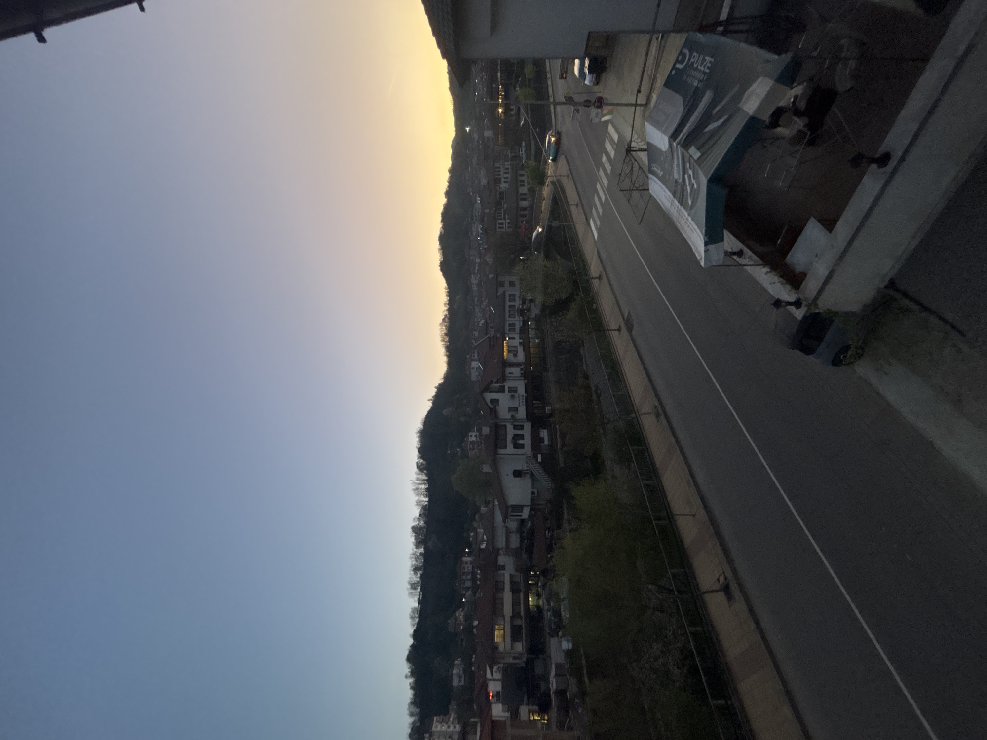 The image shows an elevated view of a section of Zlatograd, Bulgaria at dusk. The sky transitions from blue to a golden yellow on the horizon. A densely populated hillside of residential buildings stretches vertically. A road runs alongside the hillside, with crosswalks visible. A building with a "Pulze" advertisement extends into the view. The image is taken from an elevated position, potentially a balcony.
