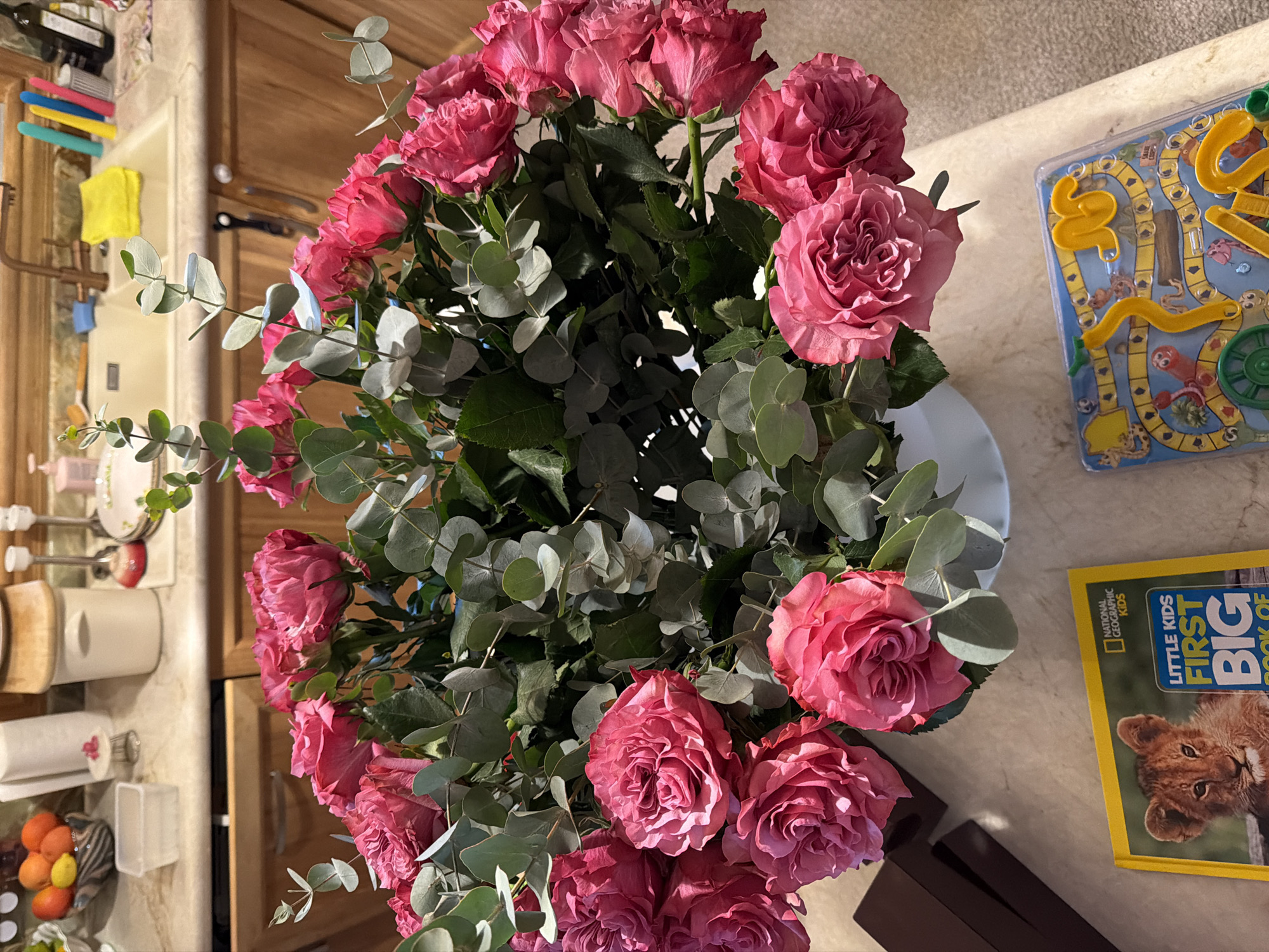The image shows a close-up view of a bouquet of pink roses and eucalyptus leaves in a white vase, placed on a marble countertop. In the background, a kitchen sink with a faucet and various kitchen items such as a yellow dishrag, colorful utensils, and a bottle of wine are visible. To the left of the vase, there are several kitchen cabinets and drawers. In the foreground, a book titled "Little Kids First Big Book of..." and a board game with animal figures and a yellow track are placed on the countertop. The overall scene appears to be a domestic setting, likely a kitchen or dining area. The location is described as Souni-Zanatzia, Cyprus.