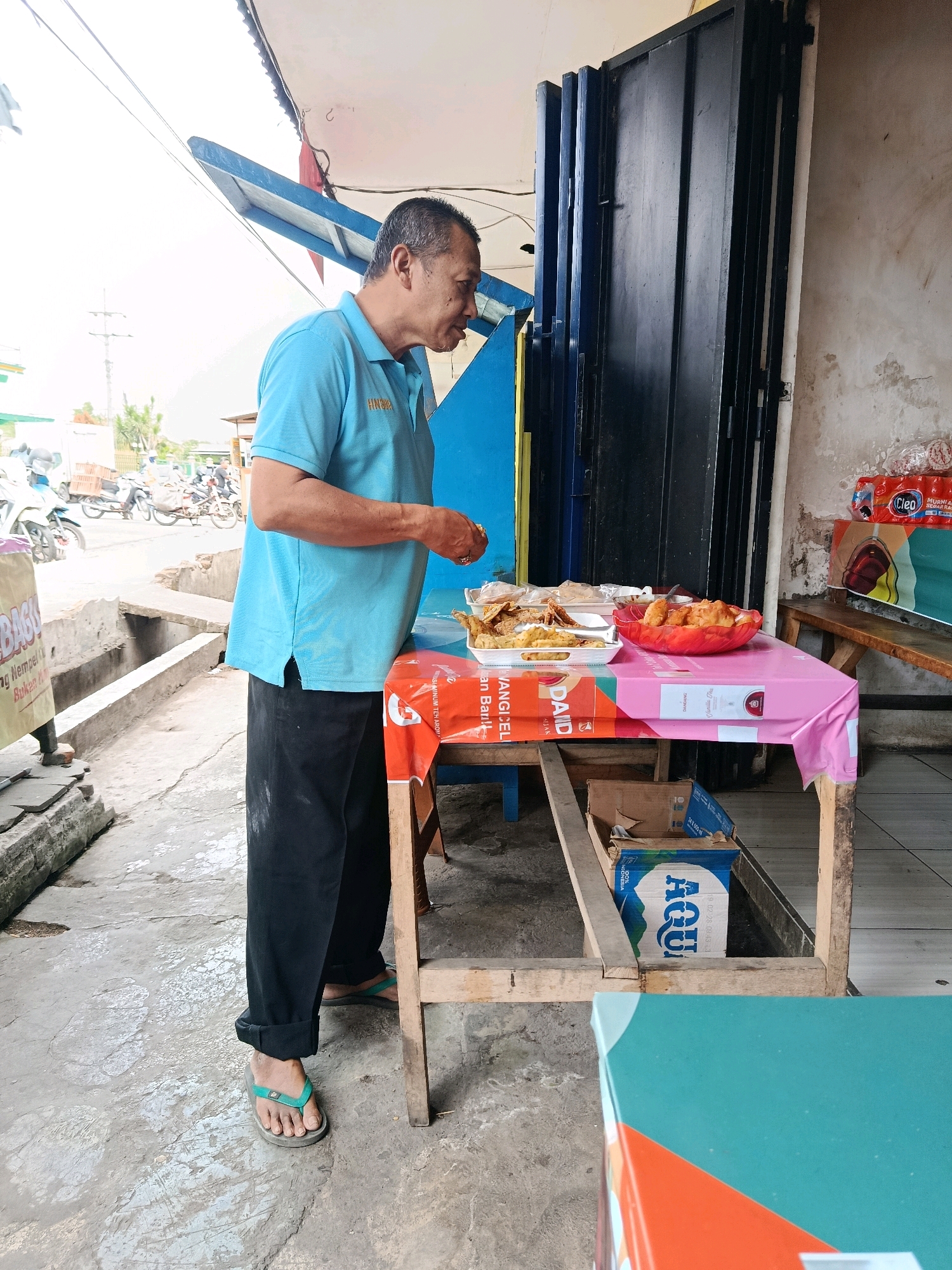 In Bululawang, Indonesia, an Asian man wearing a blue polo shirt and black pants stands at a table covered with a pink tablecloth. He is looking down at a plate of fried food. The table holds several containers of food, including fried chicken and what appears to be spring rolls. Behind the table, a dark-colored metal door is visible. To the left of the man, motorcycles are parked on a street. The ground is paved with concrete, and there are some loose bricks and a drainage channel. A cardboard box with the "Aqua" logo is placed under the table. The scene appears to be an outdoor food stall or a roadside eatery.