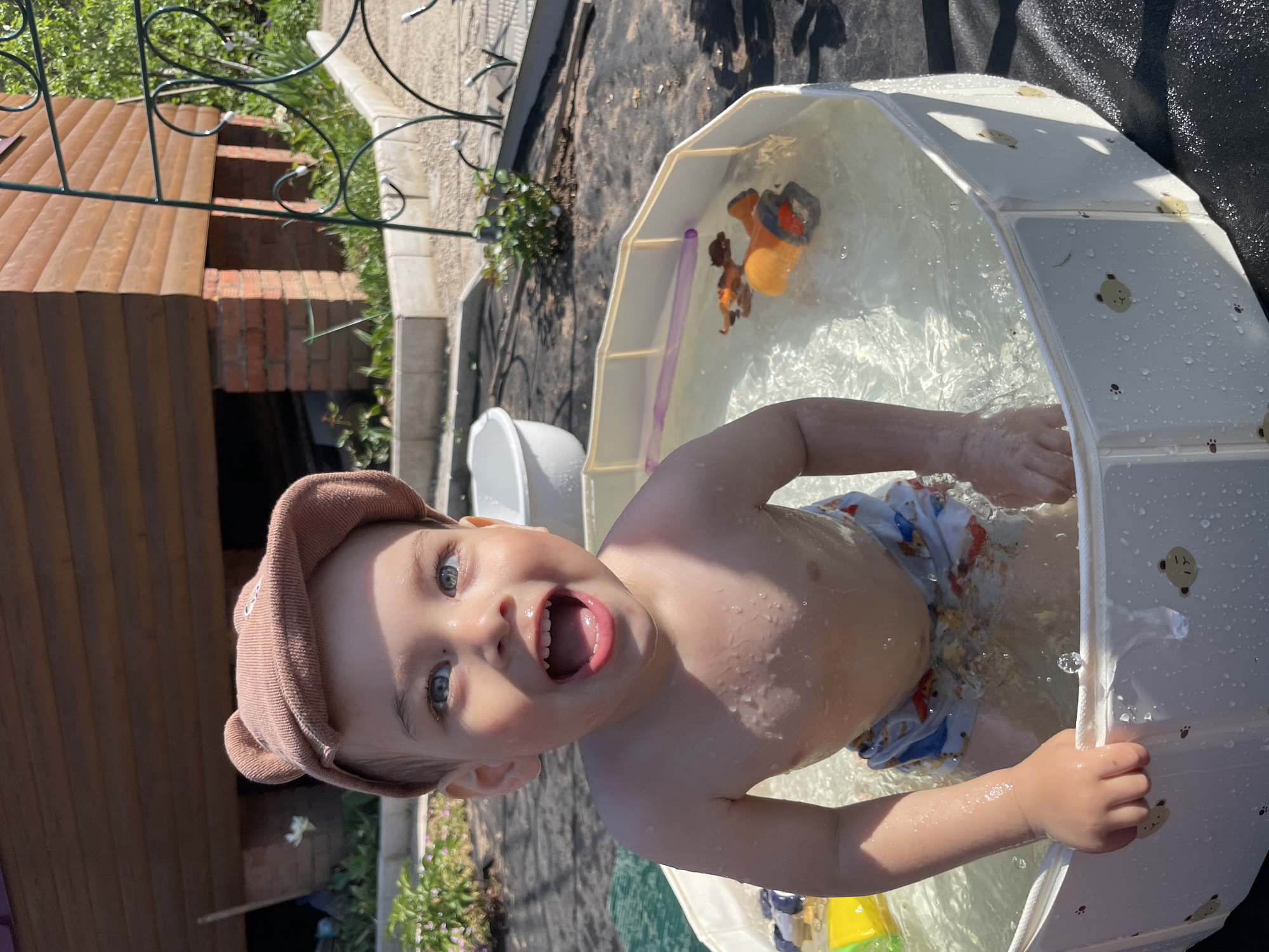 A young child, likely a toddler, stands in a white, plastic, foldable-style swimming pool filled with water. The child wears a brown cap and patterned swimming trunks. The child faces the camera with their mouth open in a possible smile or expression of excitement. A few toys are visible in the water. The pool is located outdoors, possibly in a backyard, as indicated by the presence of plants, dark ground covering, a section of brick structure with a decorative metal fence. The location is Zav'yalovo, Russia.