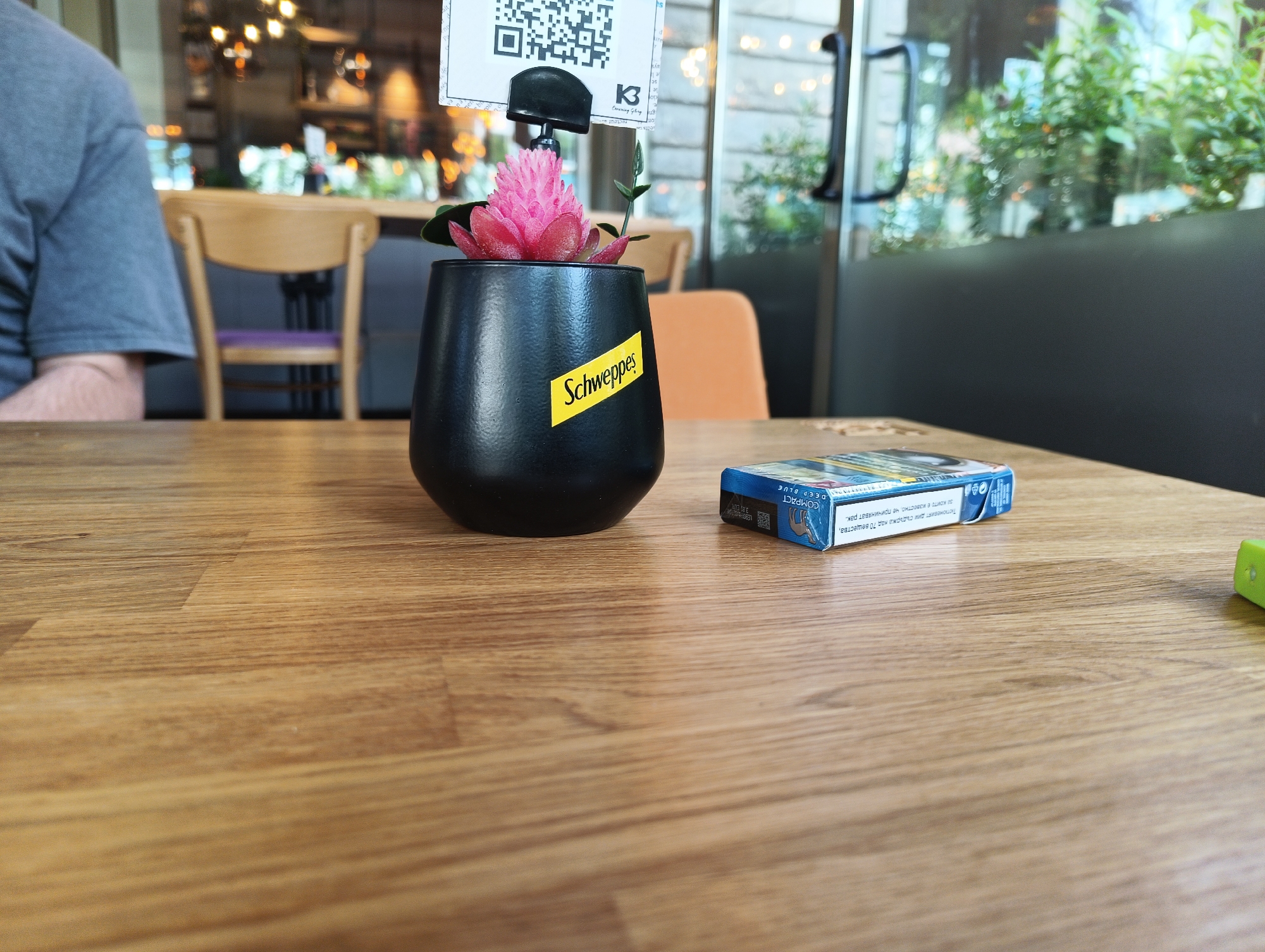 The image shows a table setting in what appears to be a restaurant or café, potentially located in Burgas, Bulgaria. A dark-haired person is partially visible on the left. On the table, there is a black pot with a faux pink flower and a menu holder displaying a QR code and the logo "K3 Crowning Georg B". Next to the pot is a pack of "Compact Deep Blue" cigarettes with Bulgarian text stating that tobacco smoke contains over 70 substances known to cause cancer. A small green object is seen at the bottom right corner of the image. The table has a light brown wood-grain surface. The background features chairs, an orange seat, lights, and what appears to be a glass door or window with plants visible on the other side. A "Schweppes" label is visible on the pot.